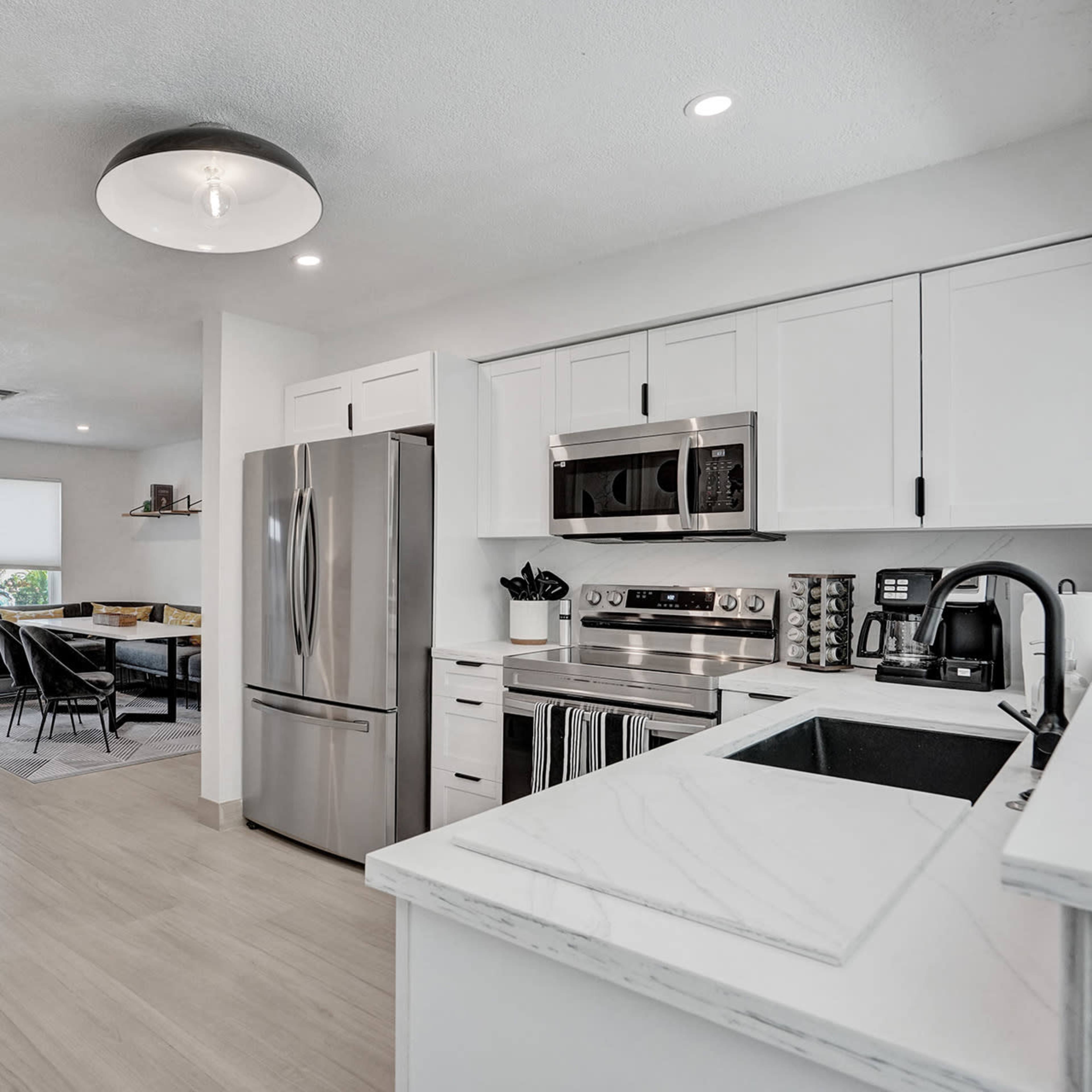 A modern kitchen with stainless steel appliances, white cabinetry, and a marble countertop, leading to a dining area with a table and chairs.