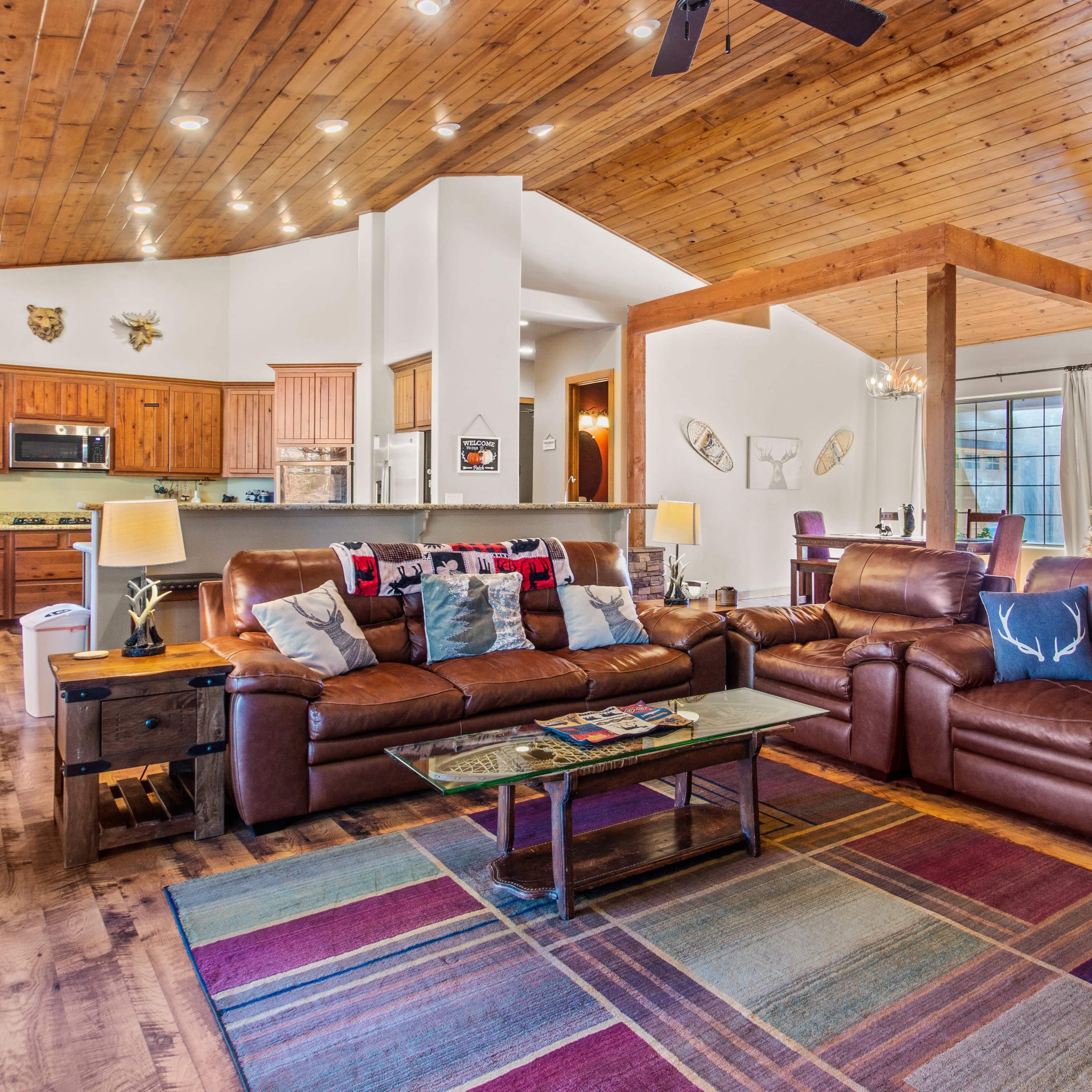 A spacious living room with two brown leather sofas, a coffee table, and a colorful area rug, surrounded by a kitchen and dining area.