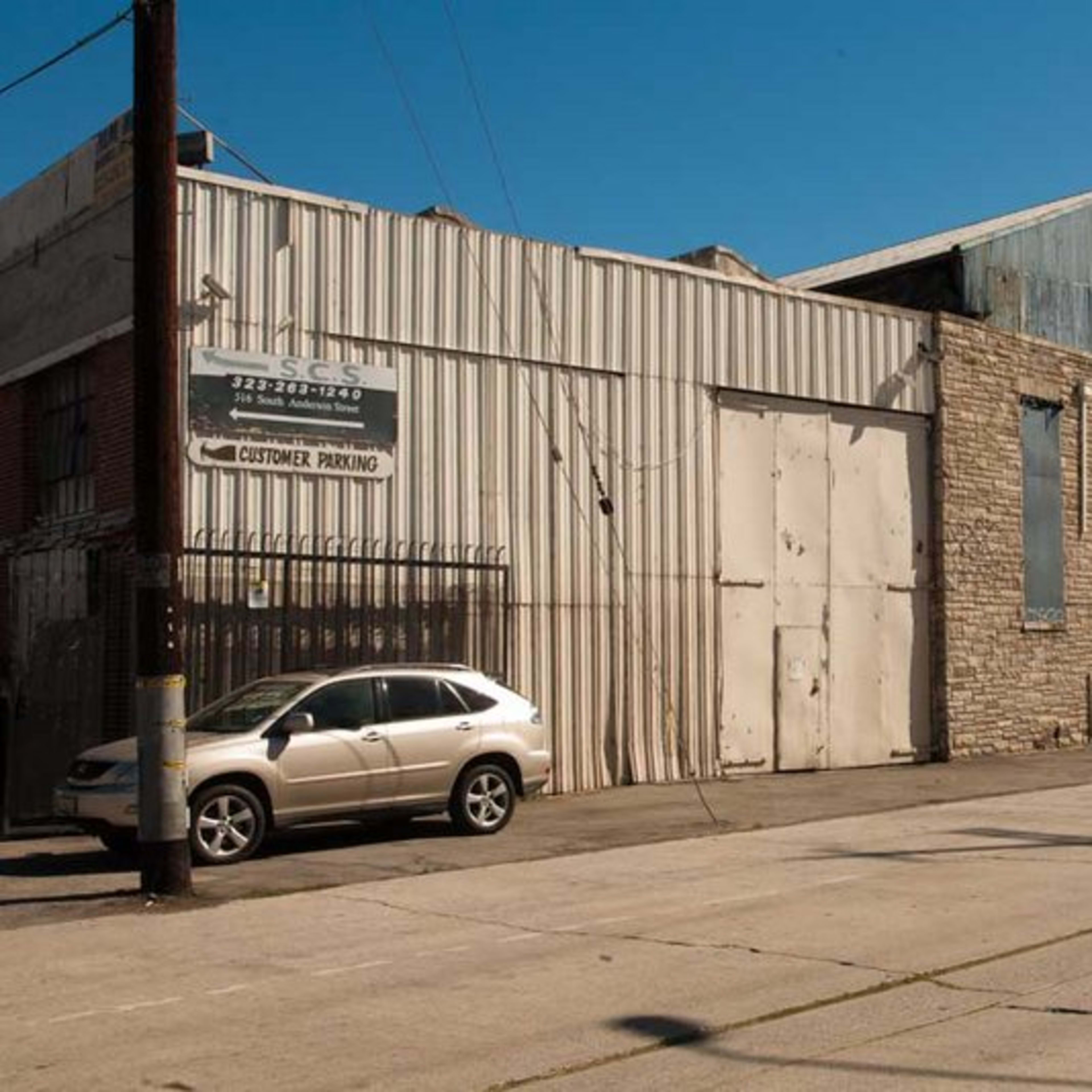 A narrow street lined with a tall, industrial building featuring metal siding and stone walls, alongside a parked silver car.
