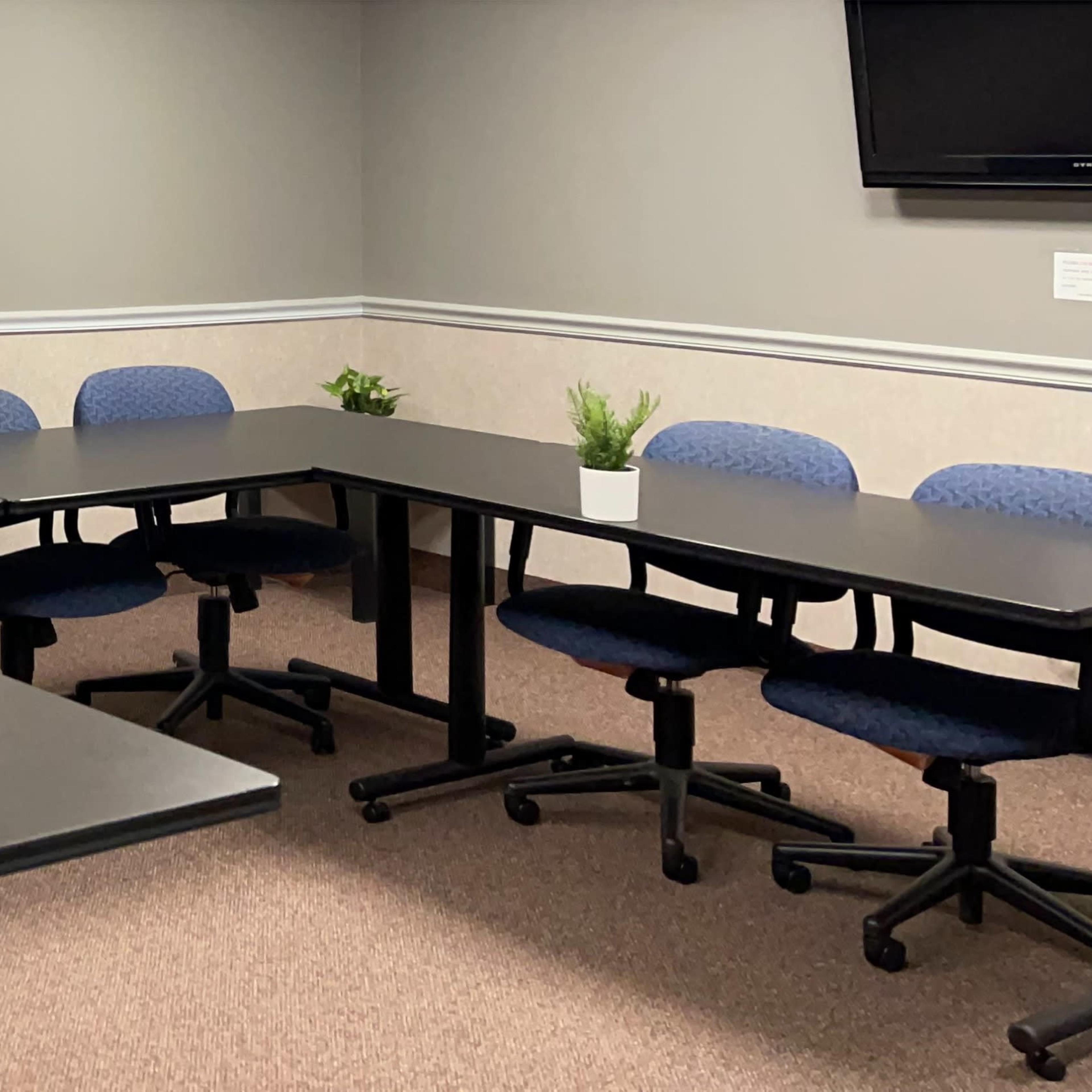 A conference room with two rectangular tables arranged in an L shape, surrounded by blue chairs and a small plant on the tables.