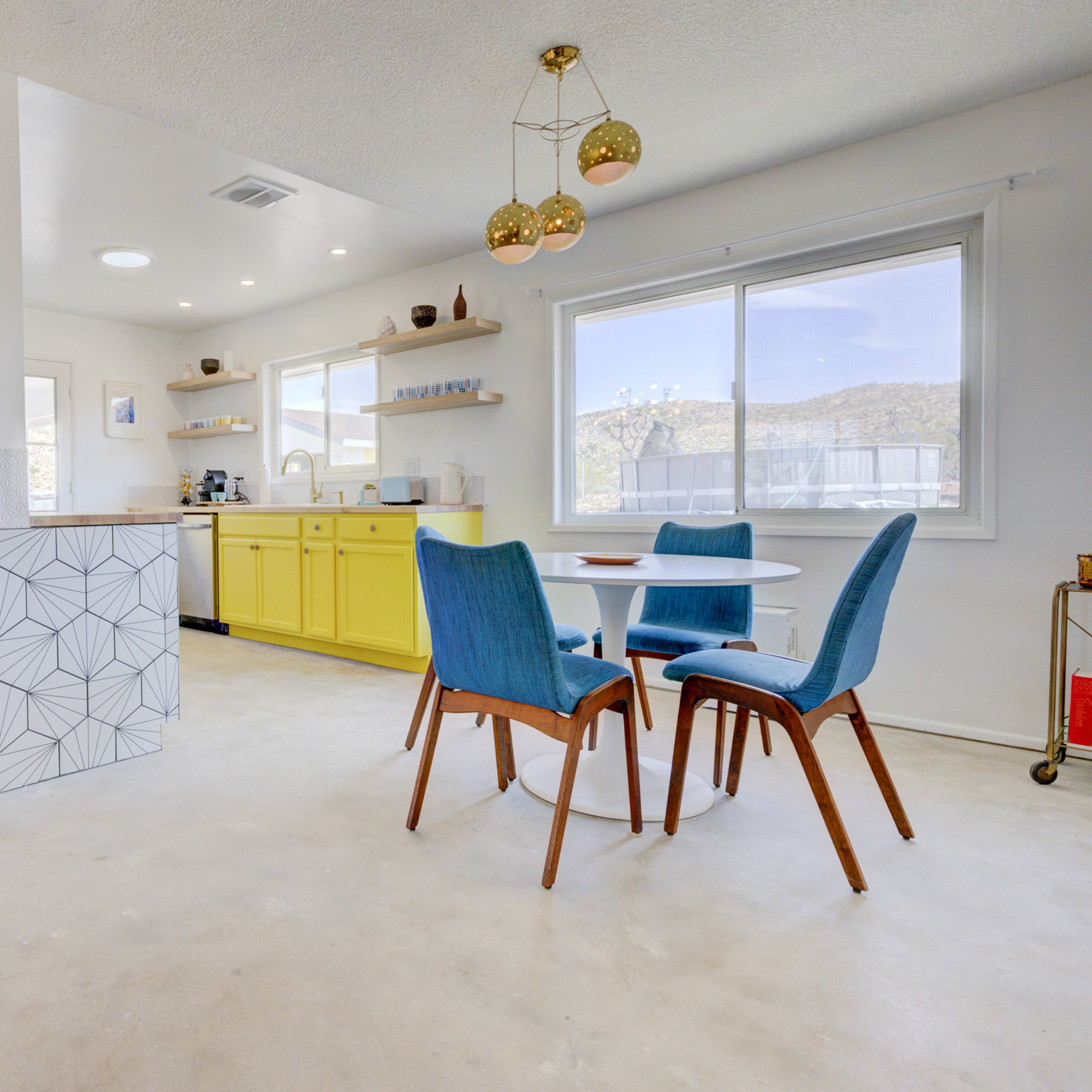 A modern kitchen and dining area features yellow cabinets, blue chairs, and a geometric-patterned bar counter, with large windows providing natural light.