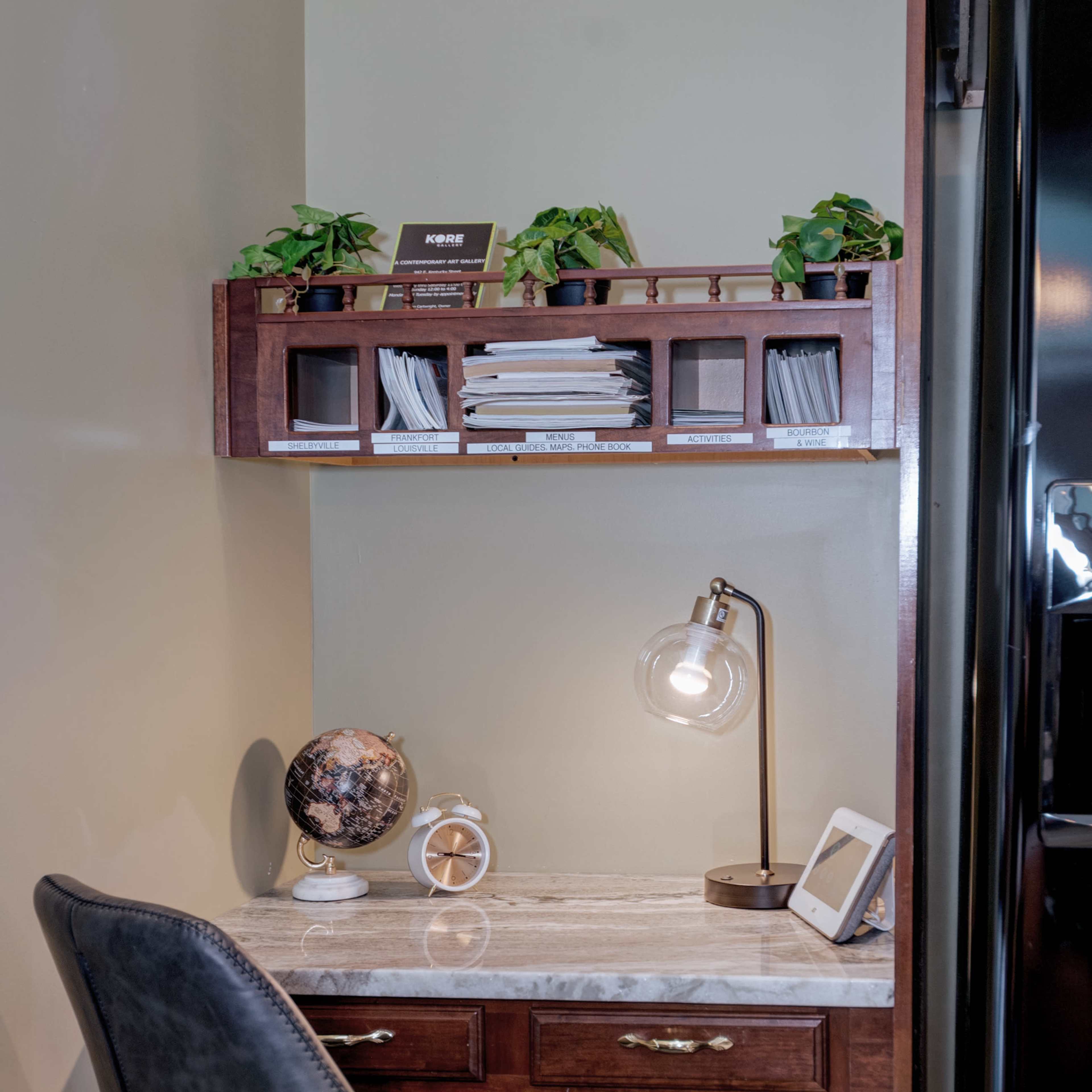 A small home office area features a desk with a marble top, a globe, a lamp, and shelves holding plants and documents against a green wall.