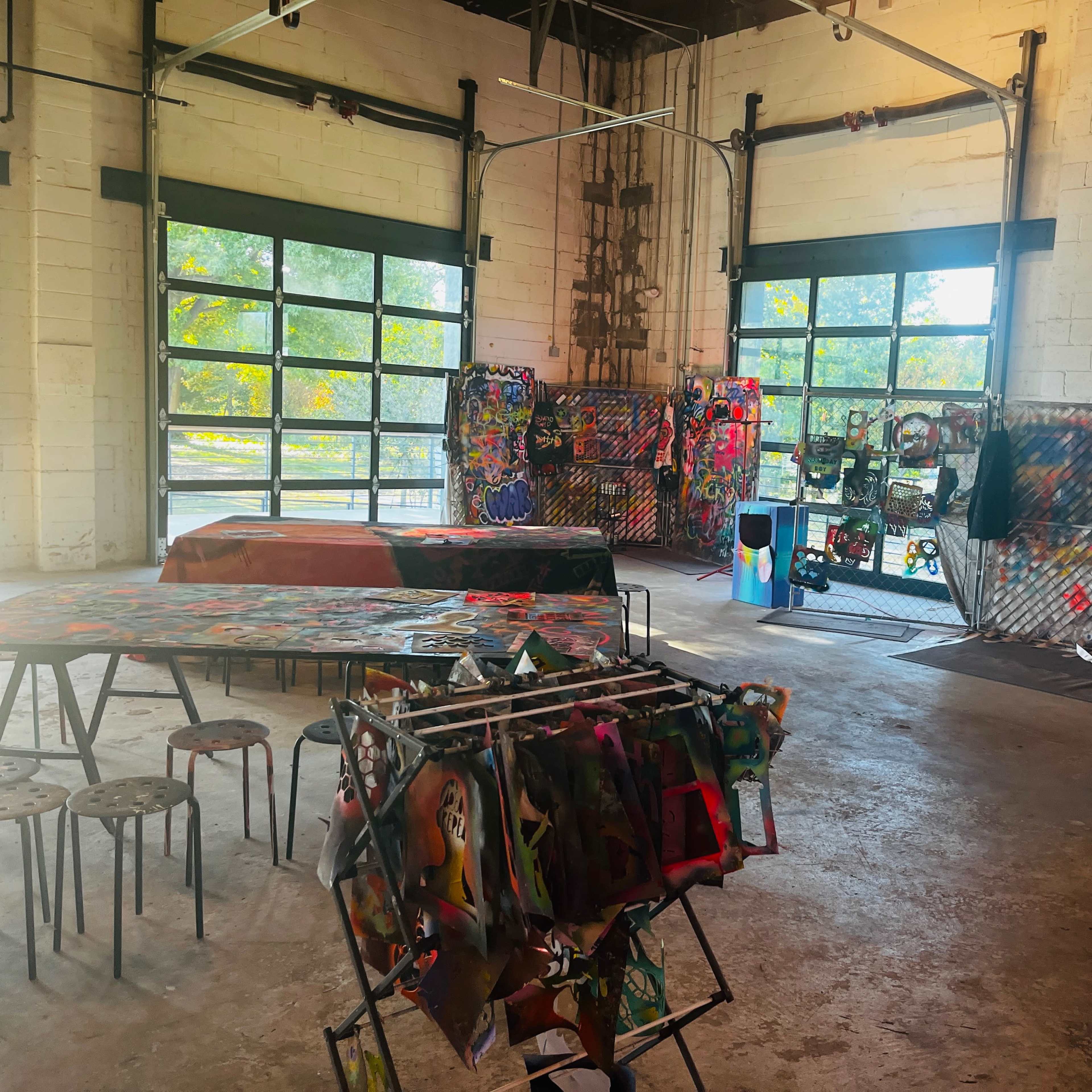 An industrial-style art space with tables displaying artwork and walls decorated with colorful pieces.