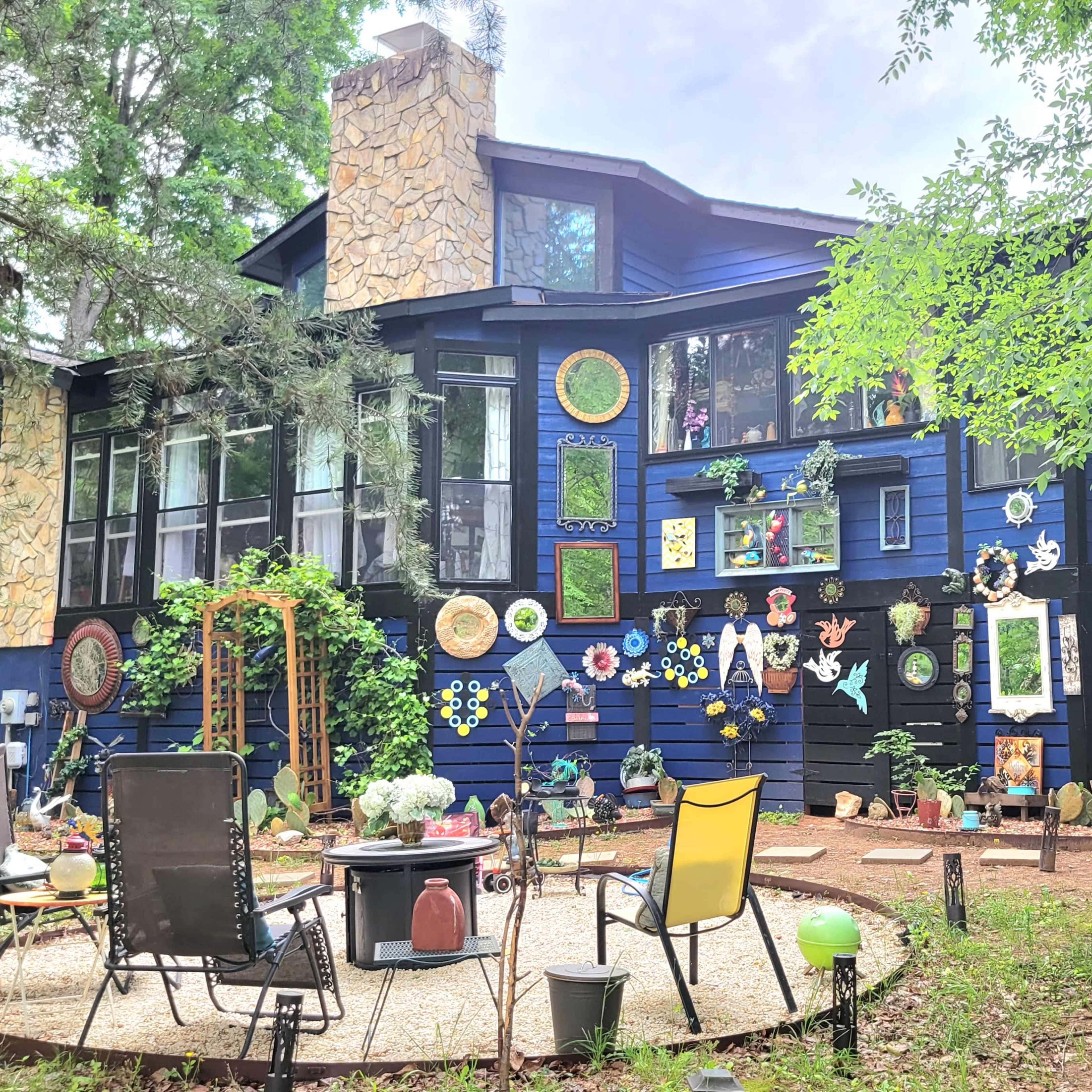 A large, dark blue house features numerous colorful decorations and art pieces on its exterior, surrounded by a circular stone patio with chairs and a fire pit.