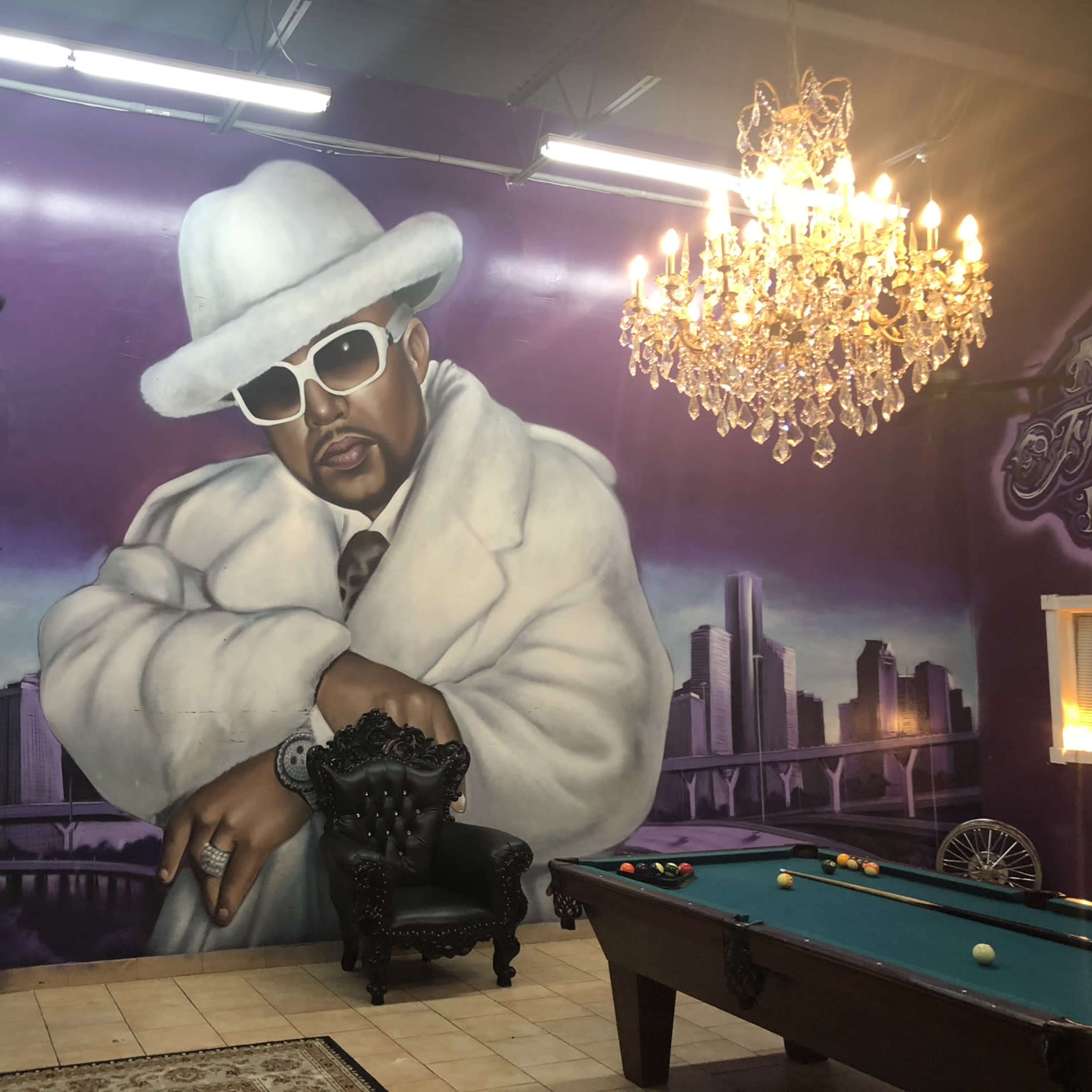 A large mural of a man in a white suit and sunglasses, set against a backdrop of a city skyline, with a pool table and chandelier in the room.