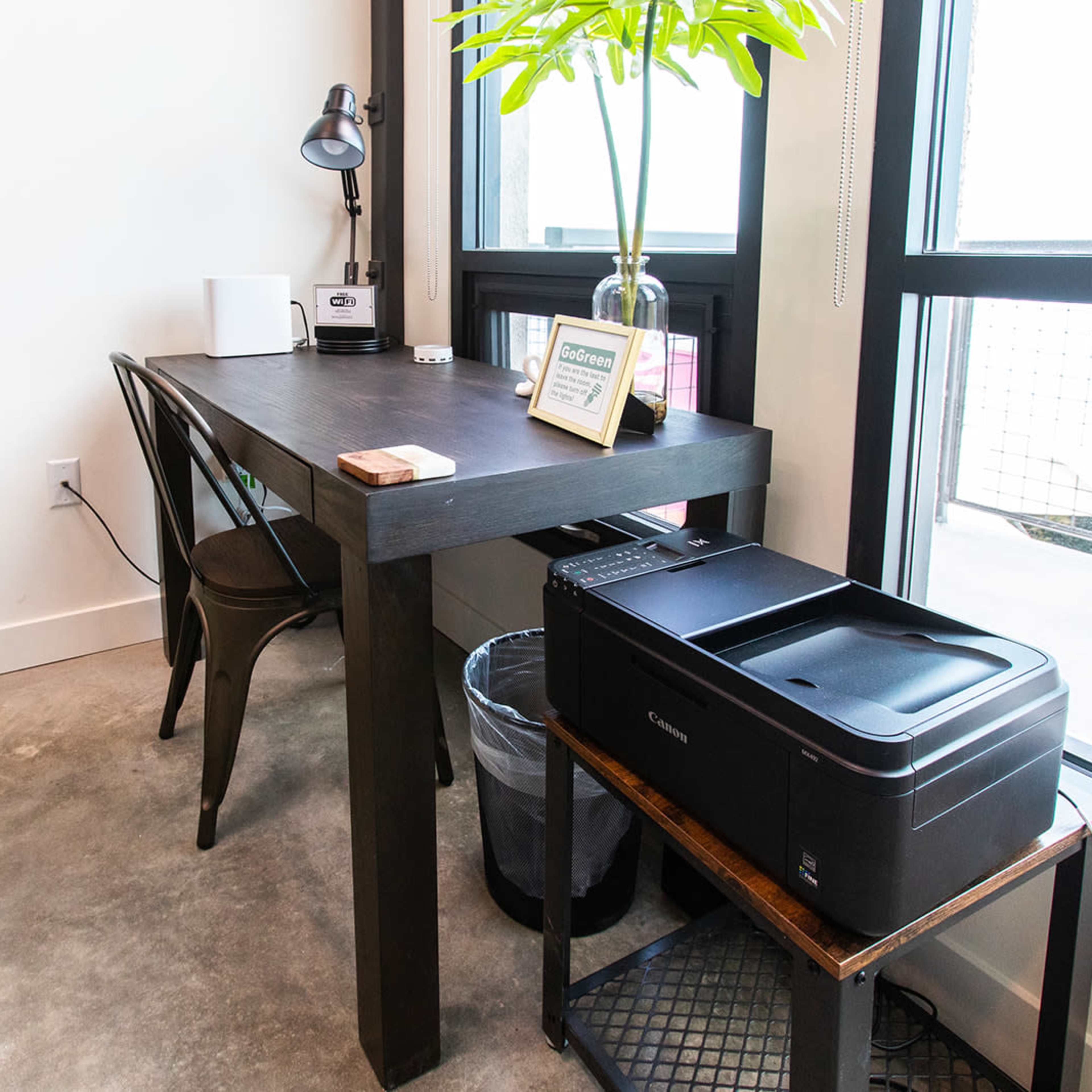 A black wooden table with a lamp, a framed picture, and a printer stands beside a window with a green accent wall in the background.