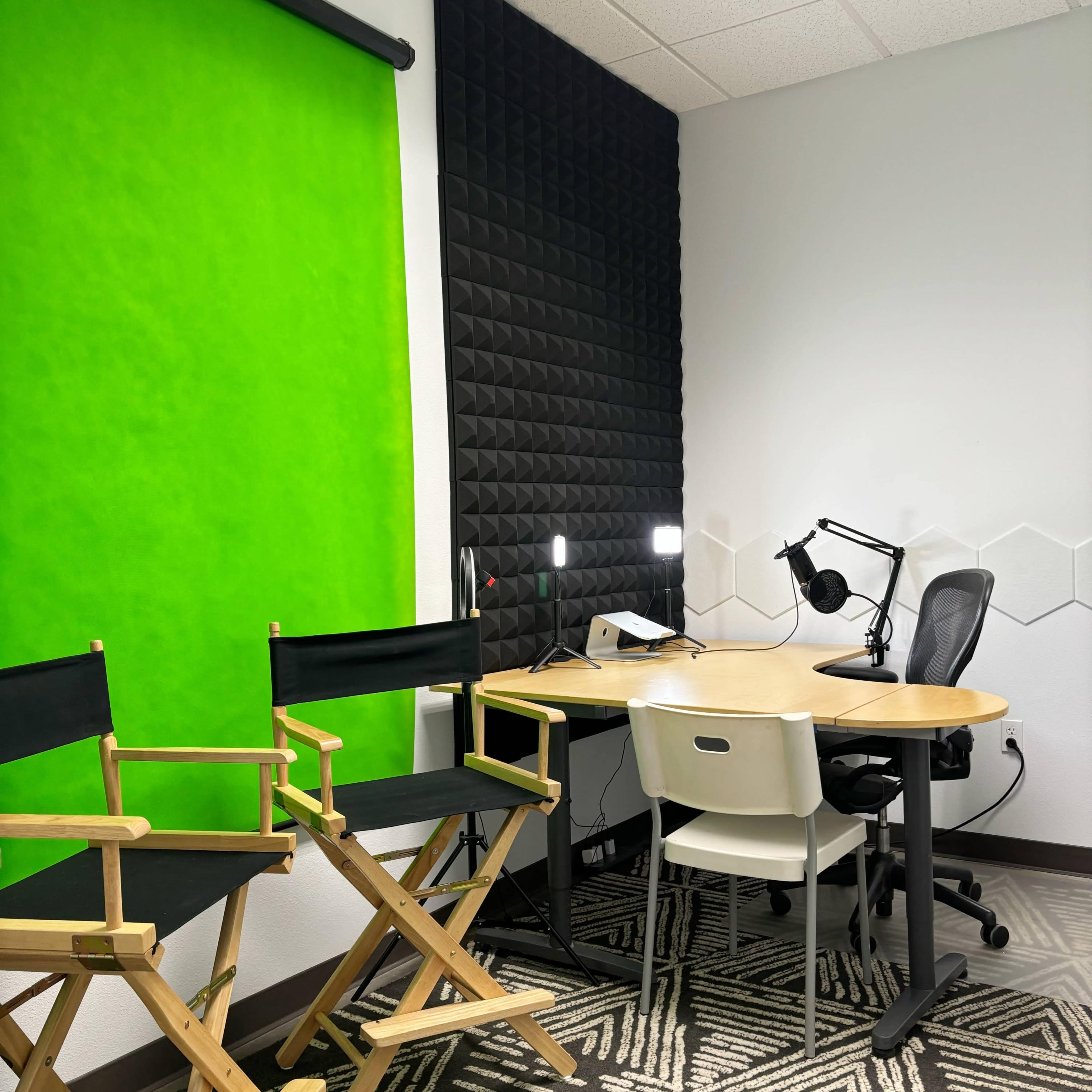 A small studio setup featuring two director's chairs, a green backdrop, a desk with a microphone attached, and acoustic paneling on one wall.