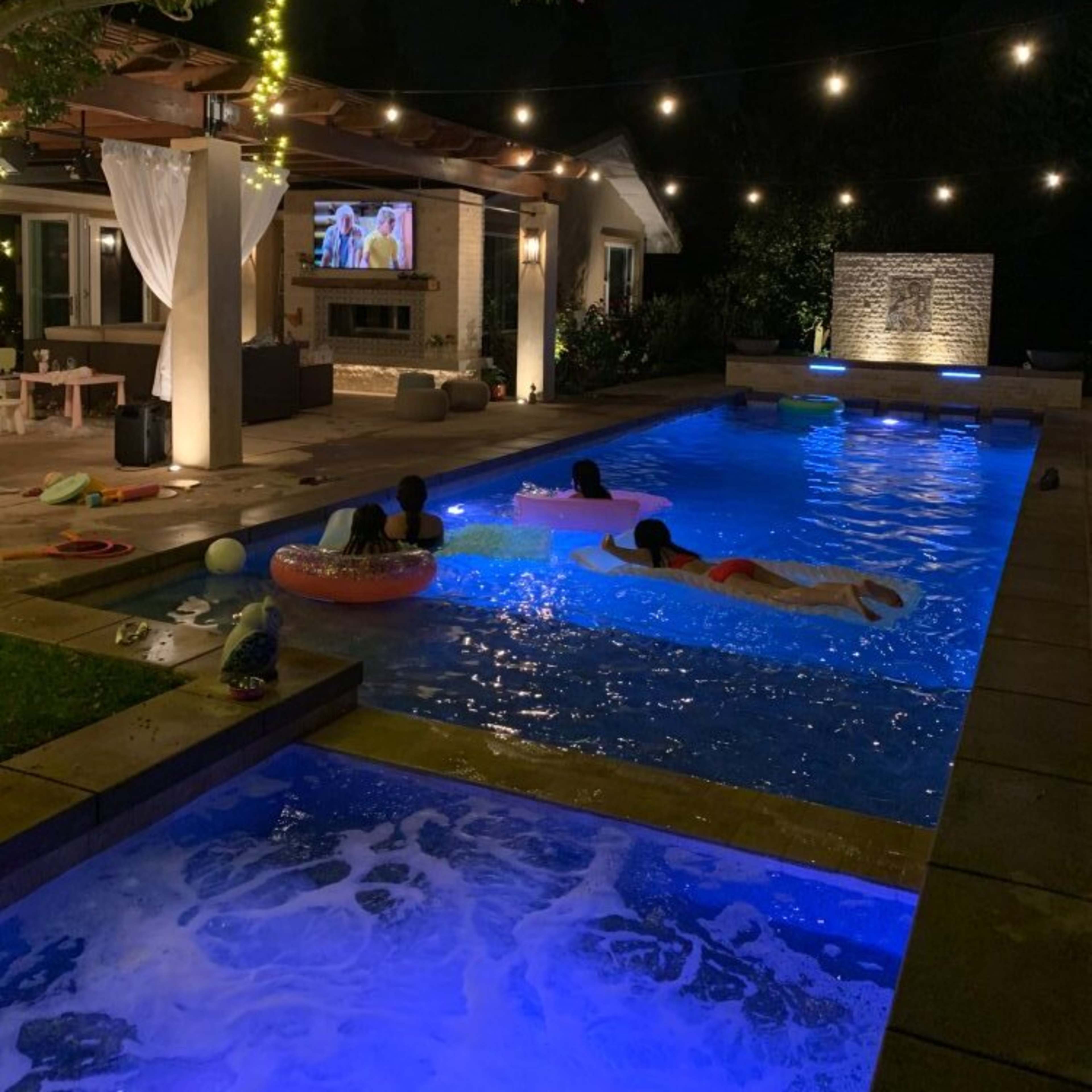 The scene depicts a backyard pool area at night, illuminated by string lights and featuring people swimming while a television displays a show nearby.
