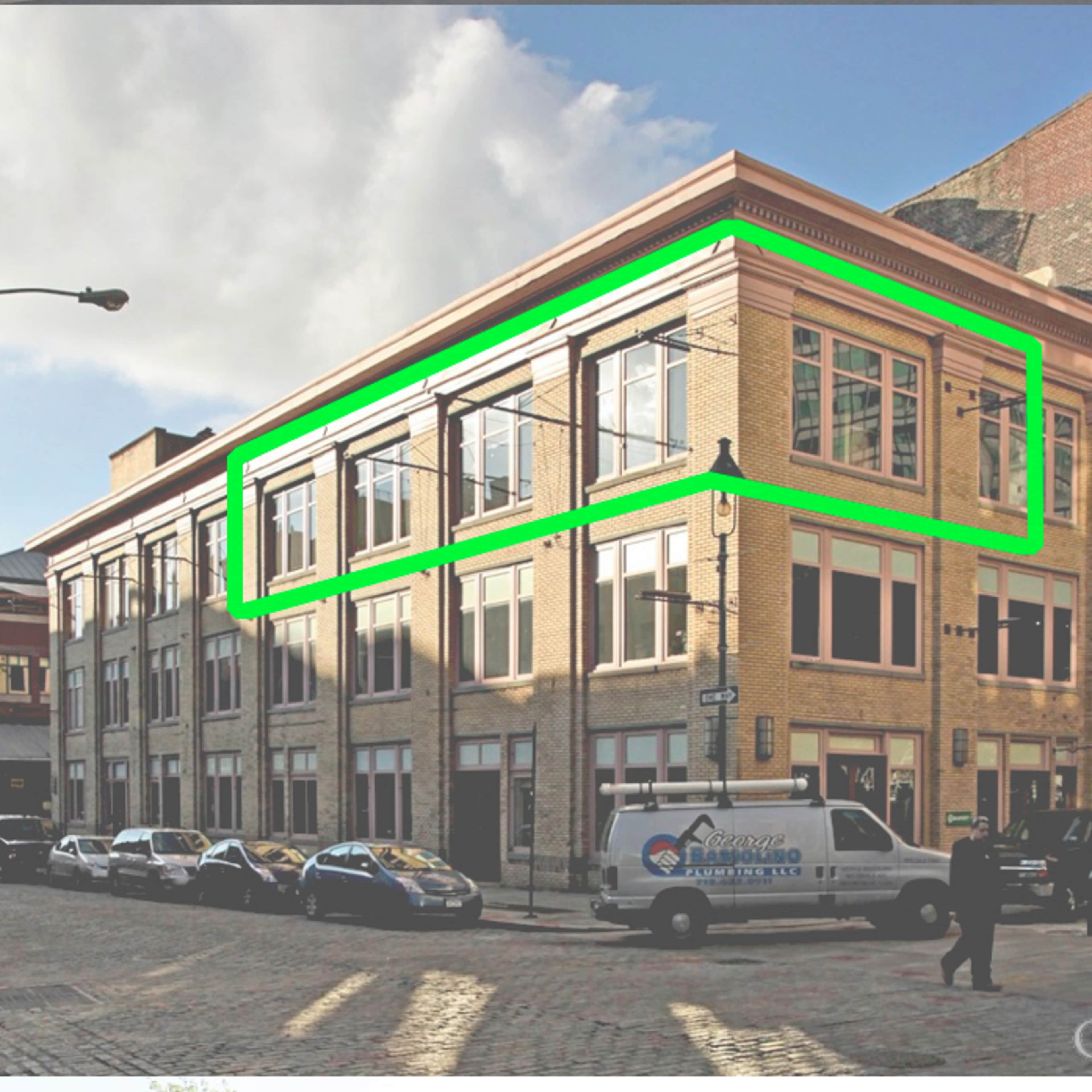 A brick building with large windows on the upper level, located on a city street with parked vehicles.