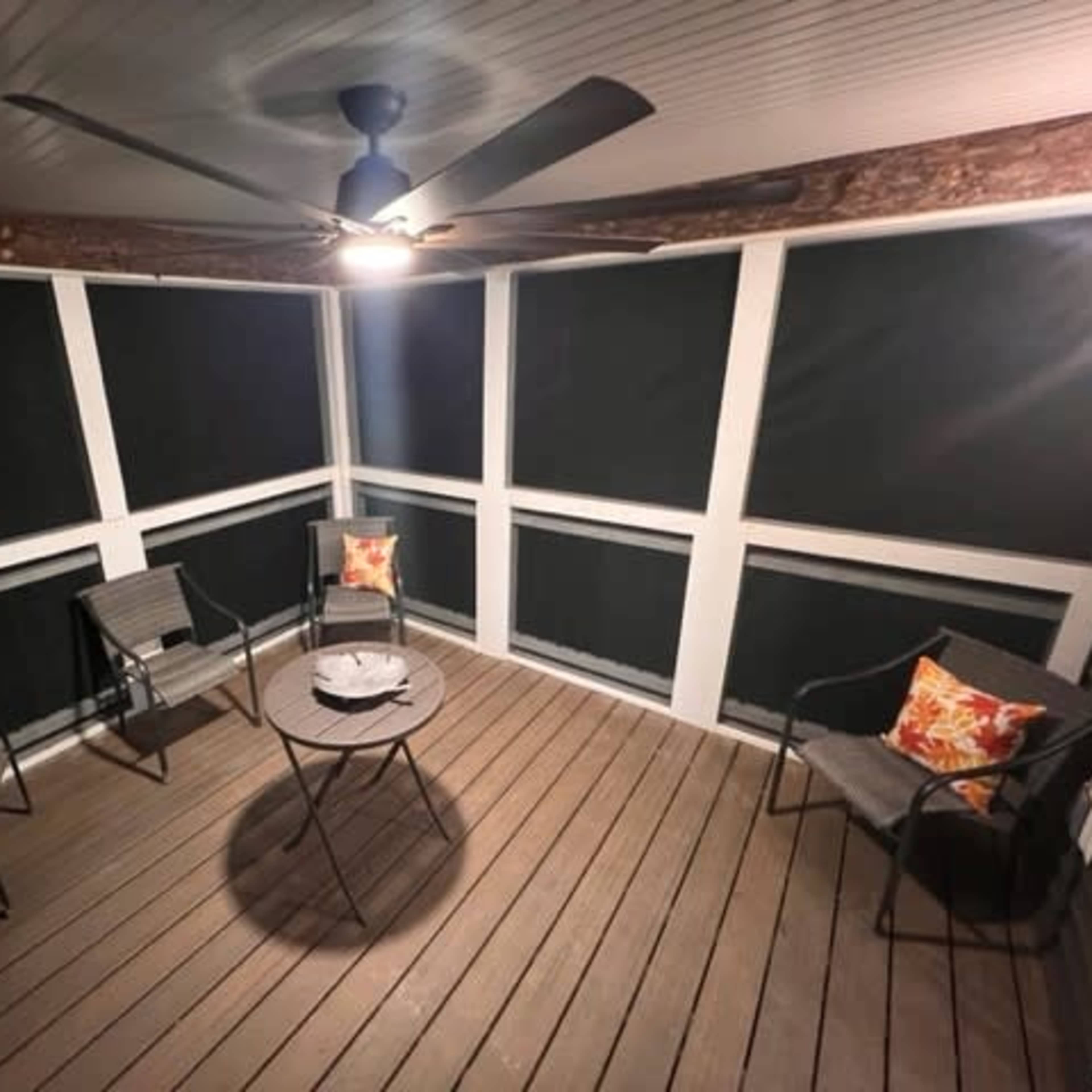 A screened-in porch features a circular table and four chairs, with decorative pillows, under a ceiling fan.