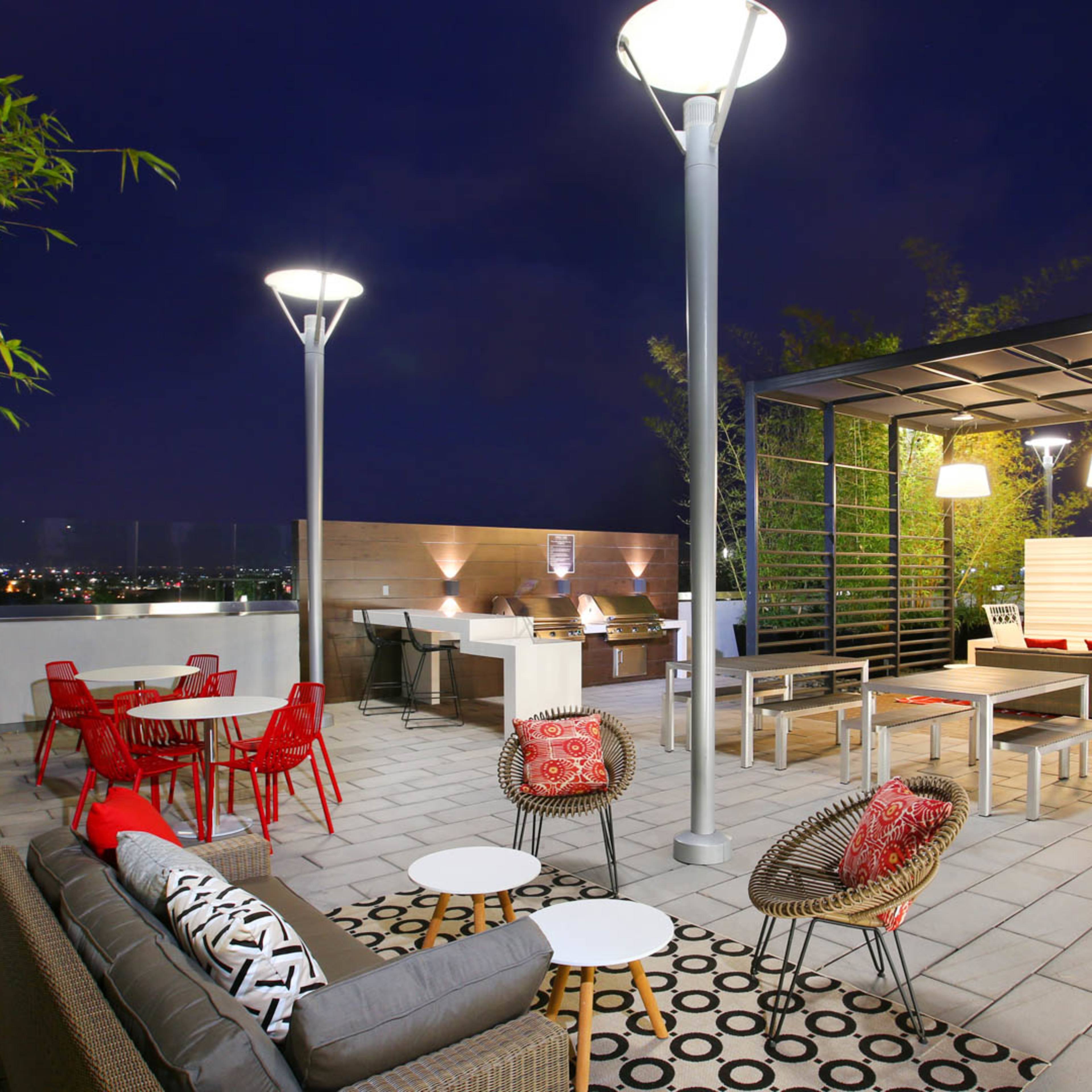 A modern rooftop patio illuminated by lamps, featuring a mix of seating areas, tables, and greenery against a night skyline.