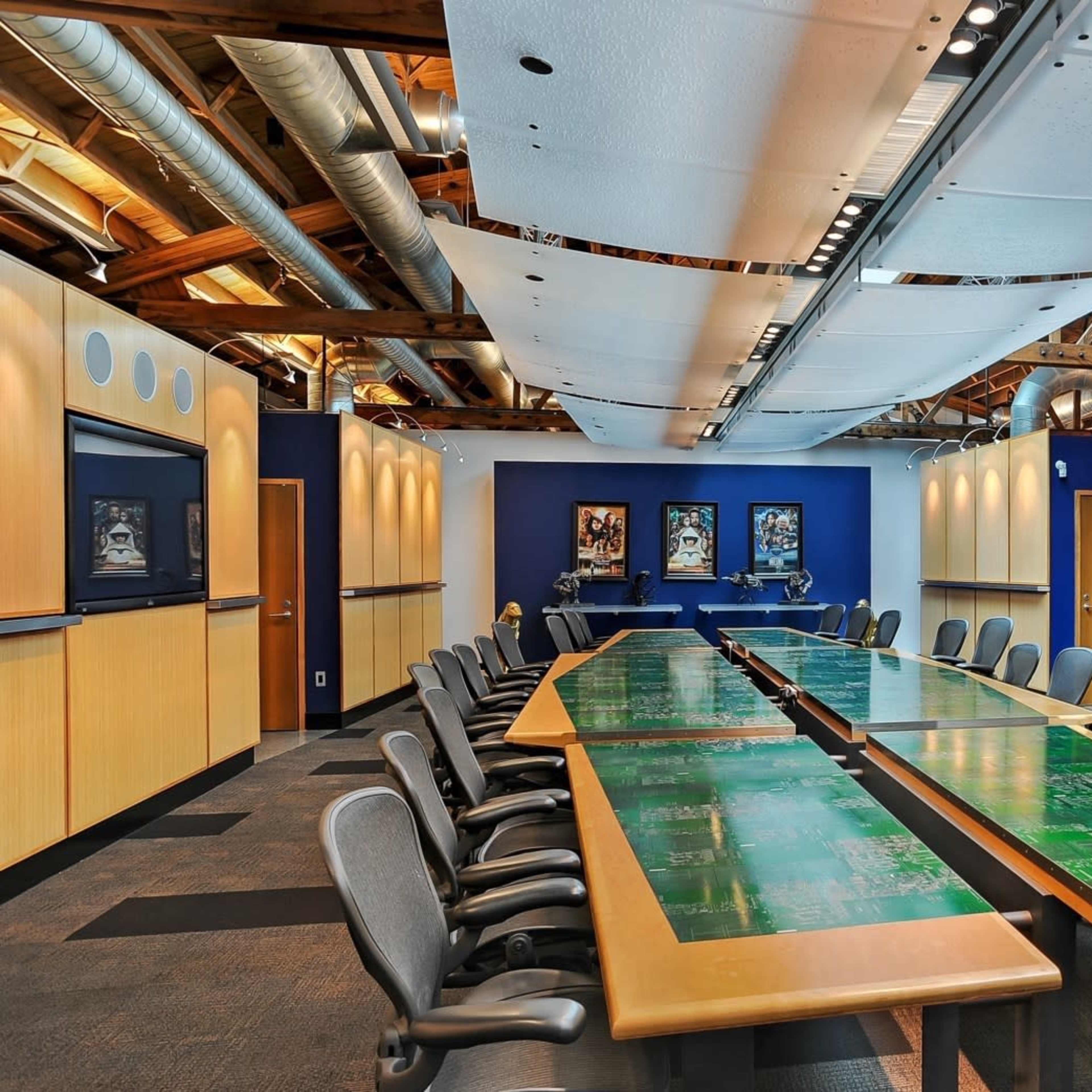 A spacious conference room features a long table with a green surface, surrounded by ergonomic chairs and equipped with large screens on the walls.