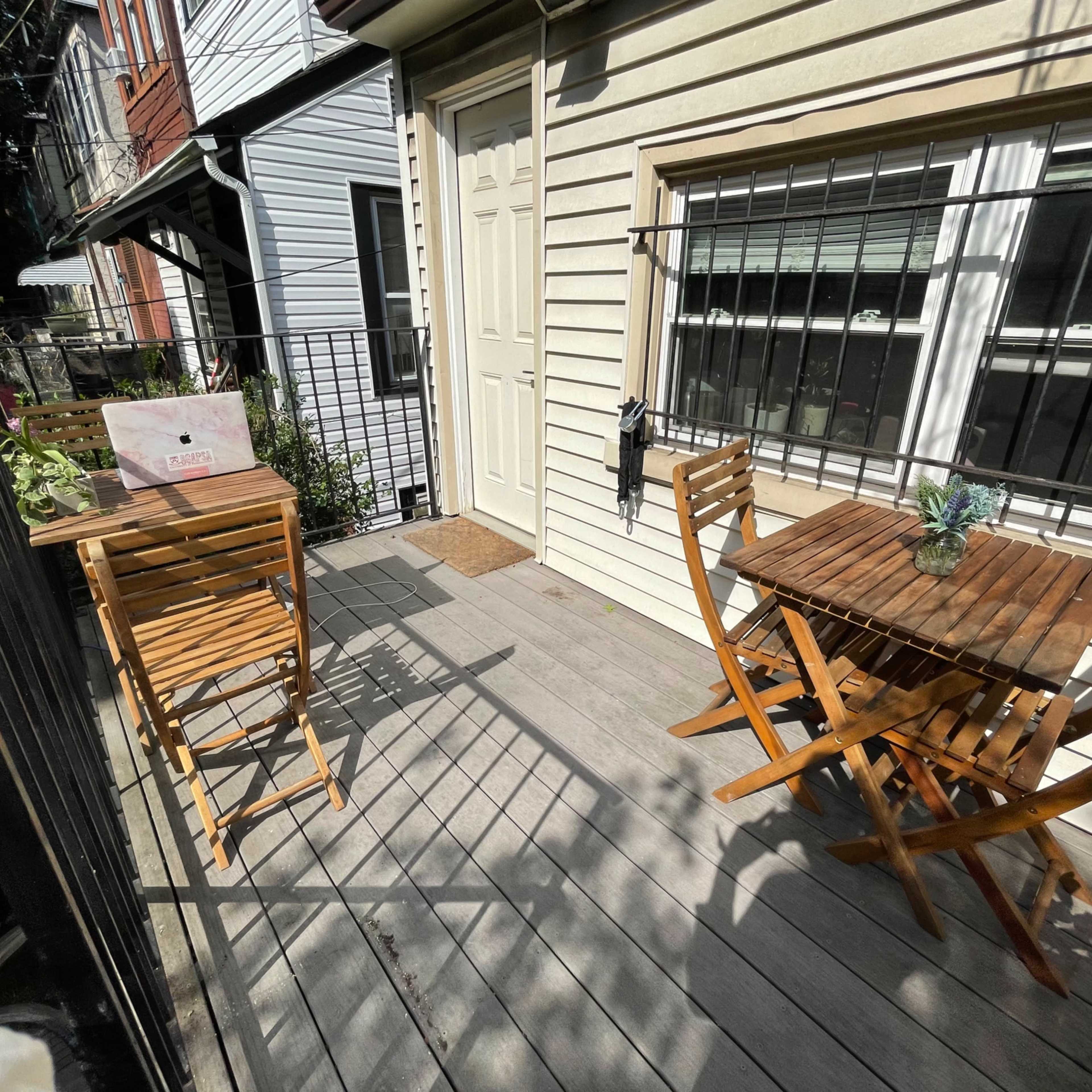A small balcony with two wooden tables and chairs, surrounded by buildings and greenery.