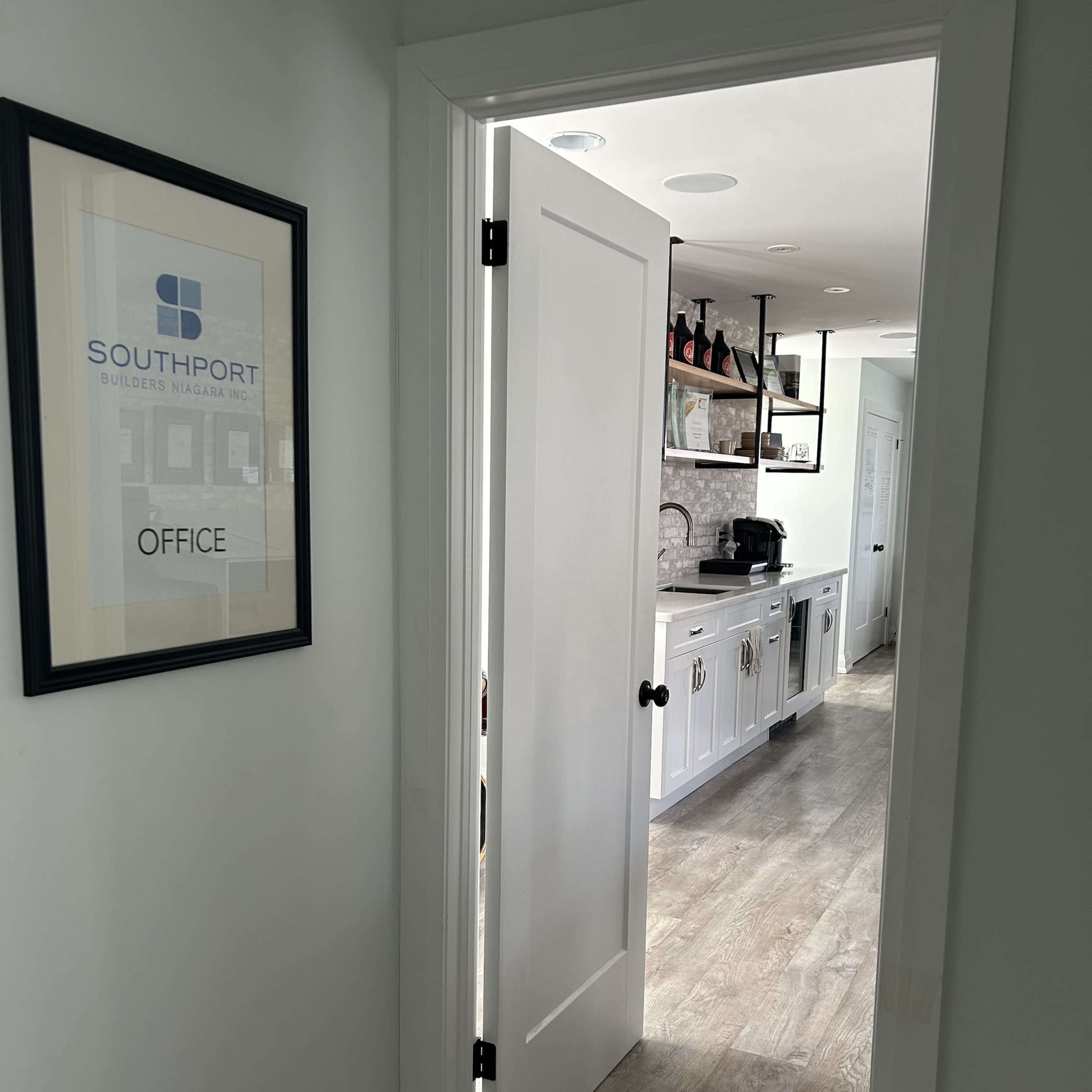 A partially open door leads to a well-lit office space with a view of a kitchen featuring cabinets and an espresso machine.