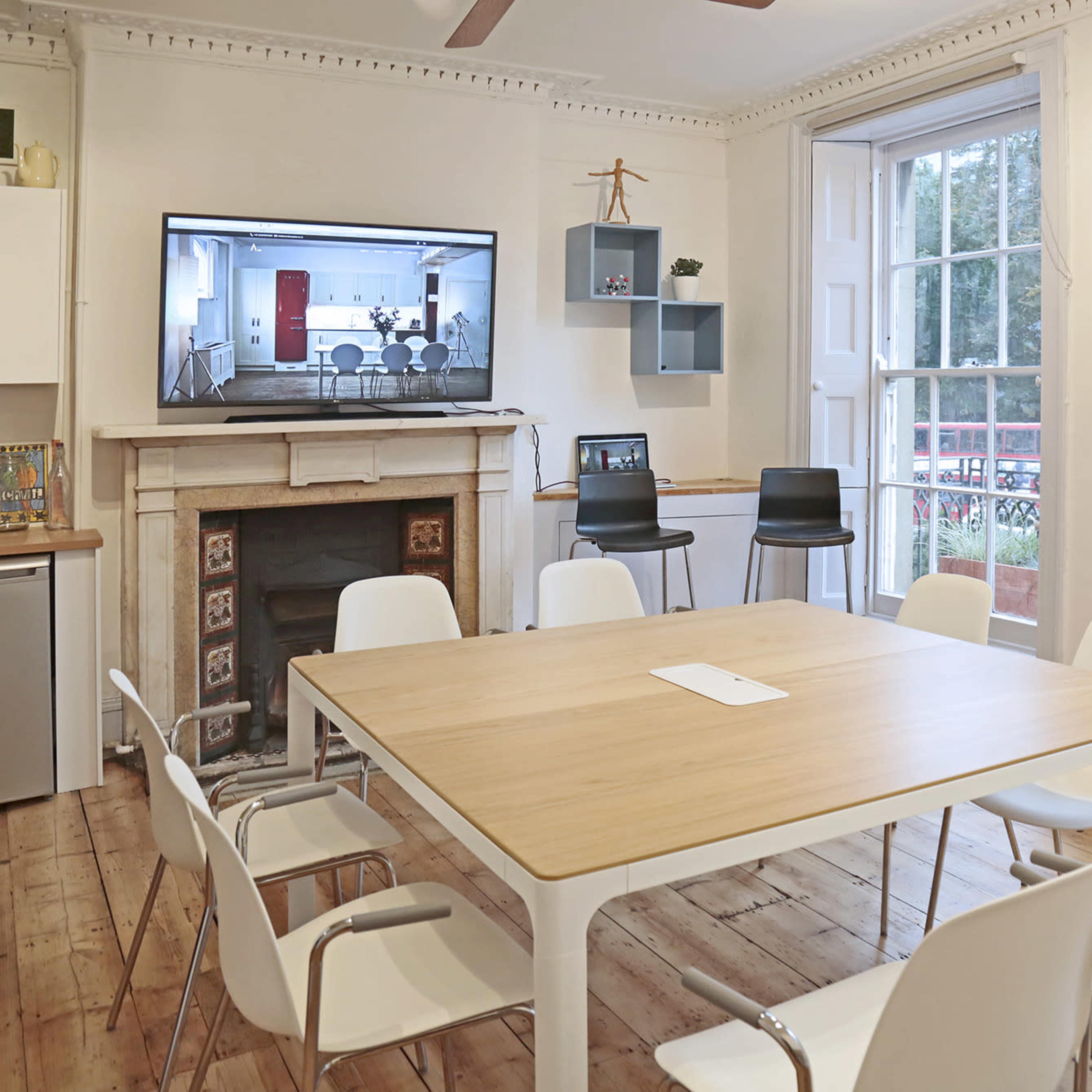 A dining area features a wooden table surrounded by white chairs, a wall-mounted television displaying a meeting room, and large windows with a view.