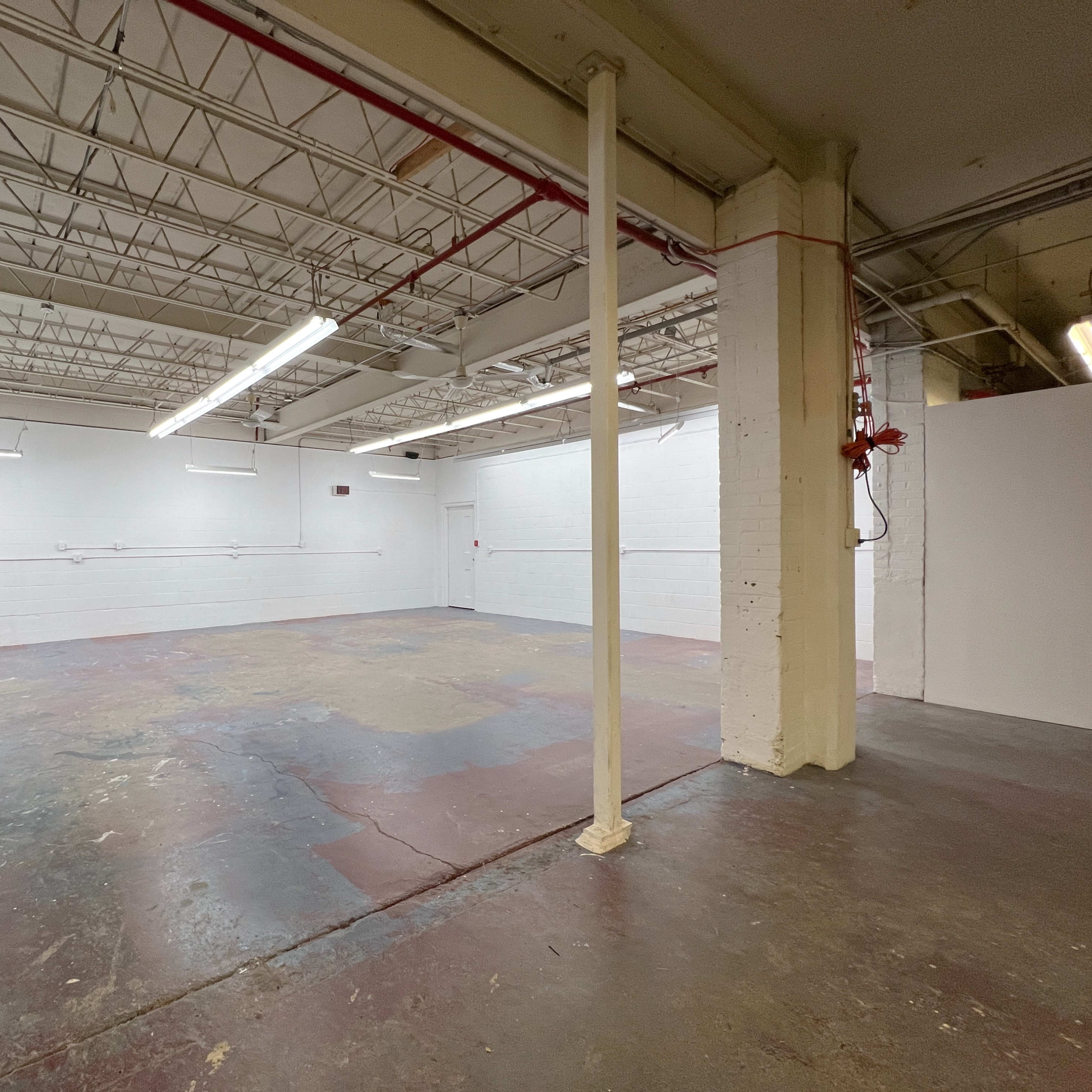 A spacious, empty room with white walls and a concrete floor, featuring exposed pipes and a column in the center.