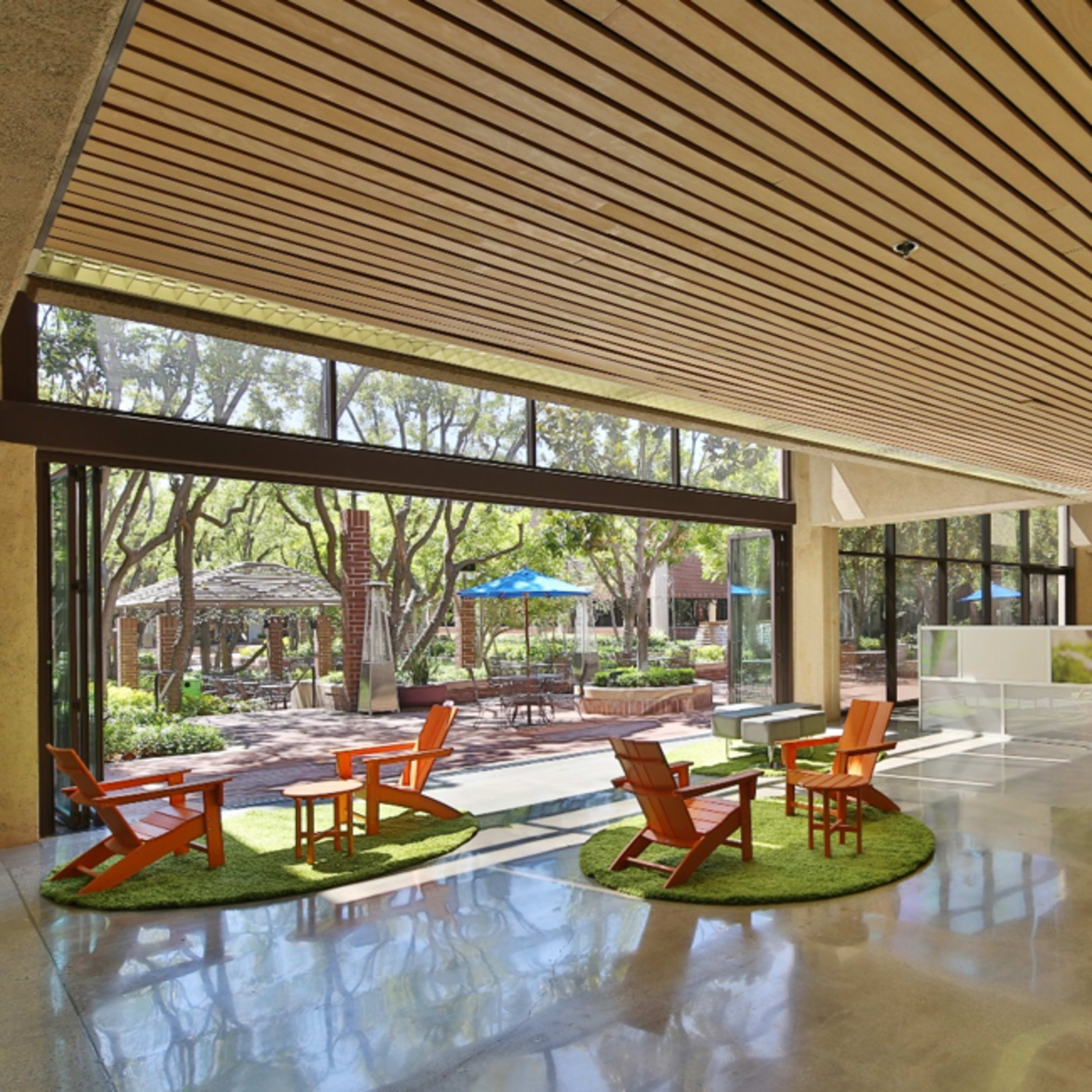 A modern interior space featuring wooden beams, a smooth floor, and orange chairs arranged on green rugs, with large windows offering views of an outdoor area with trees and seating.
