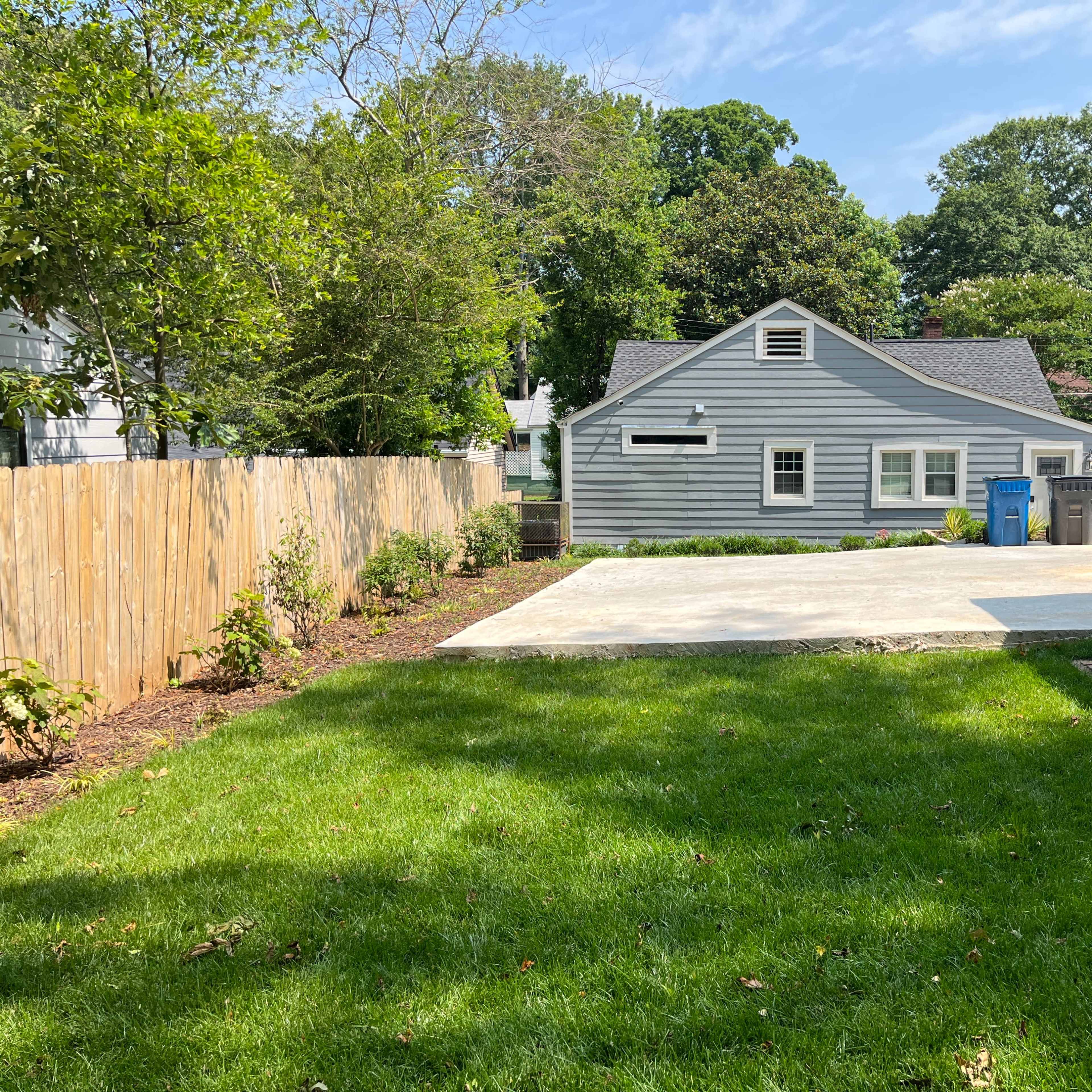 A grassy backyard with a concrete patio area, bordered by a wooden fence and featuring a small building in the background.