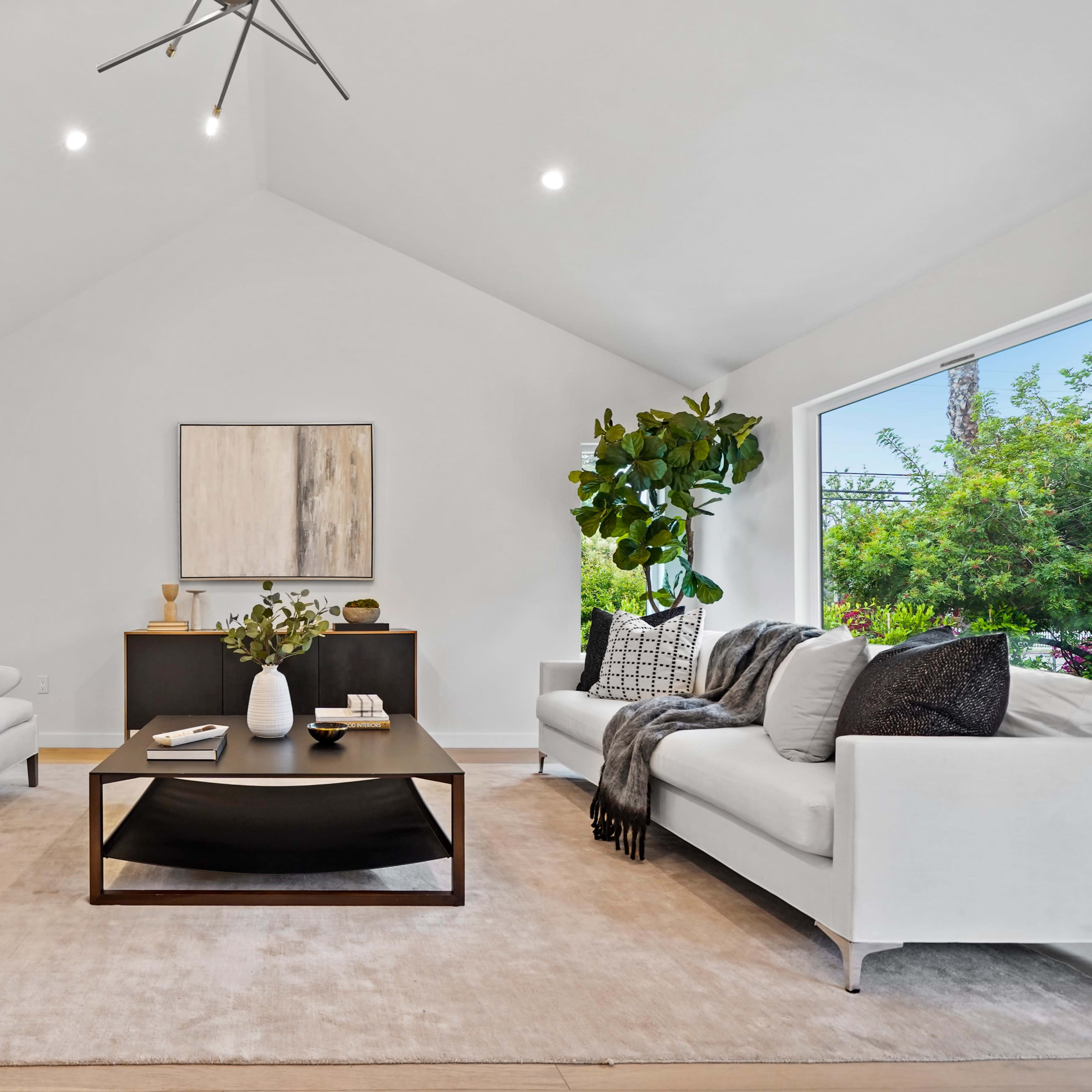 A modern living room features a black coffee table at the center, surrounded by two white armchairs and a white sofa against neutral walls and large windows.