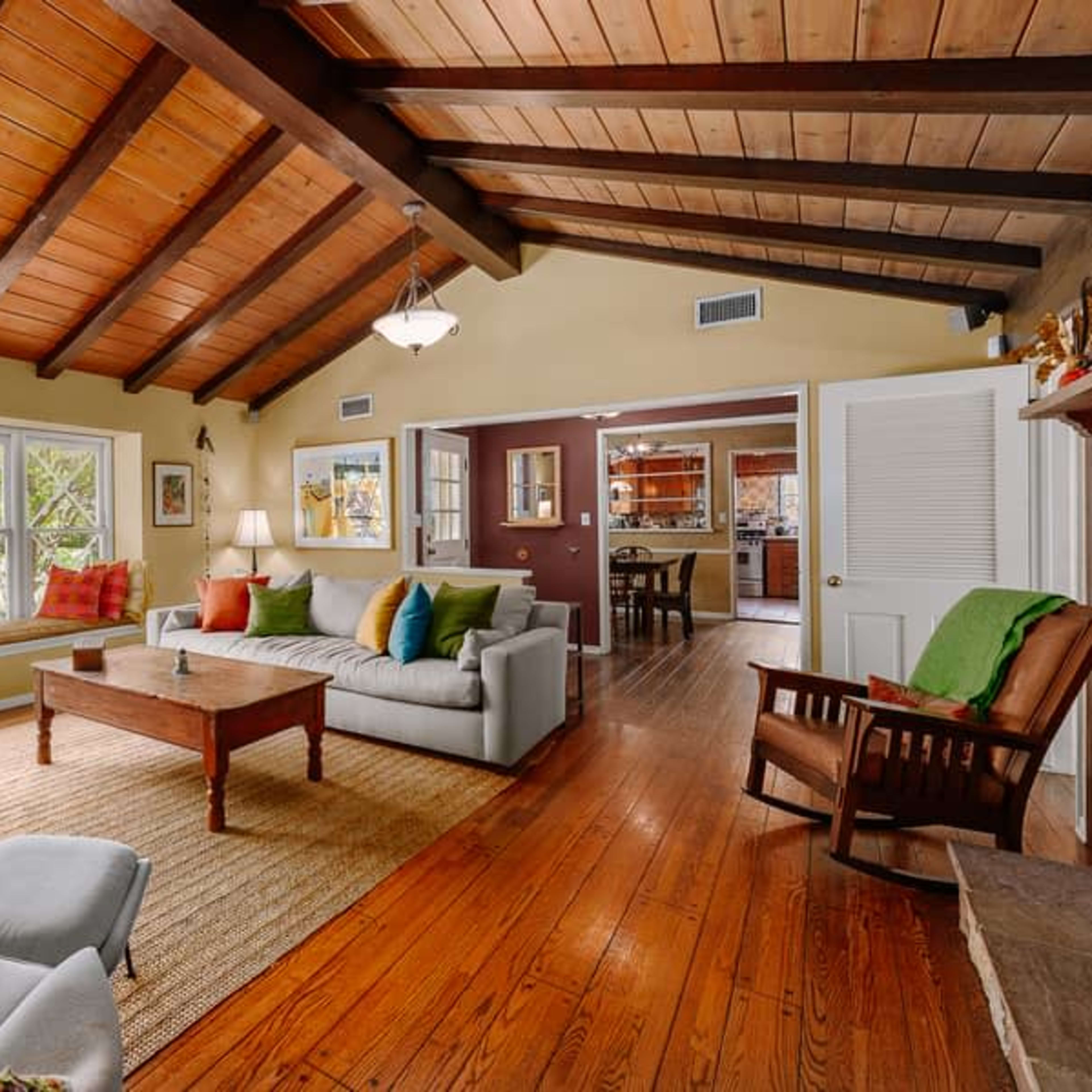A cozy living room with a wooden beam ceiling, a large window facing a garden, and a mix of seating options including a couch and rocking chair.