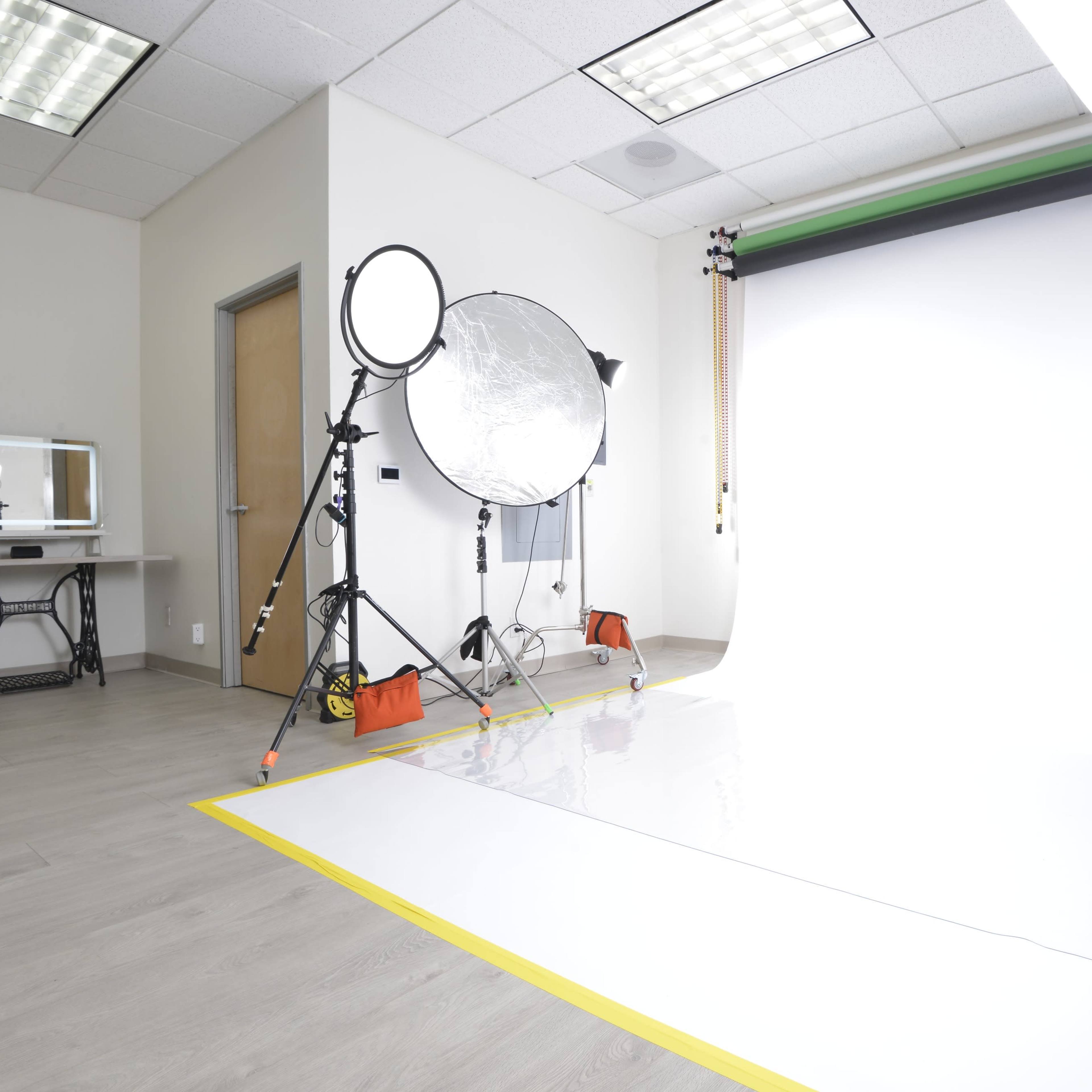A photography studio equipped with lighting equipment, a reflective umbrella, and a white backdrop, along with a rolling cart and makeup station.