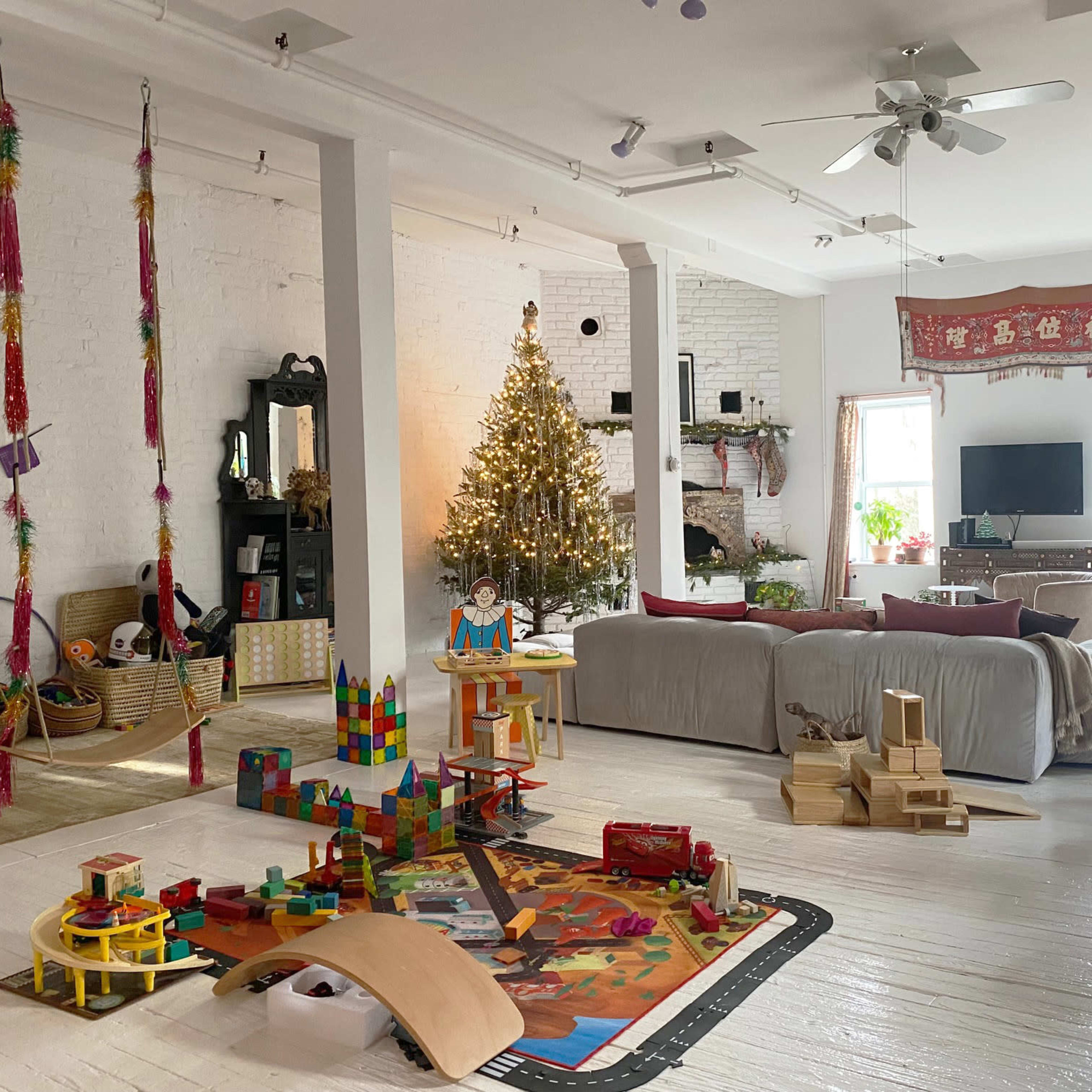 A bright living room features a Christmas tree, a play area with toys, and a cozy seating arrangement with a television in the background.