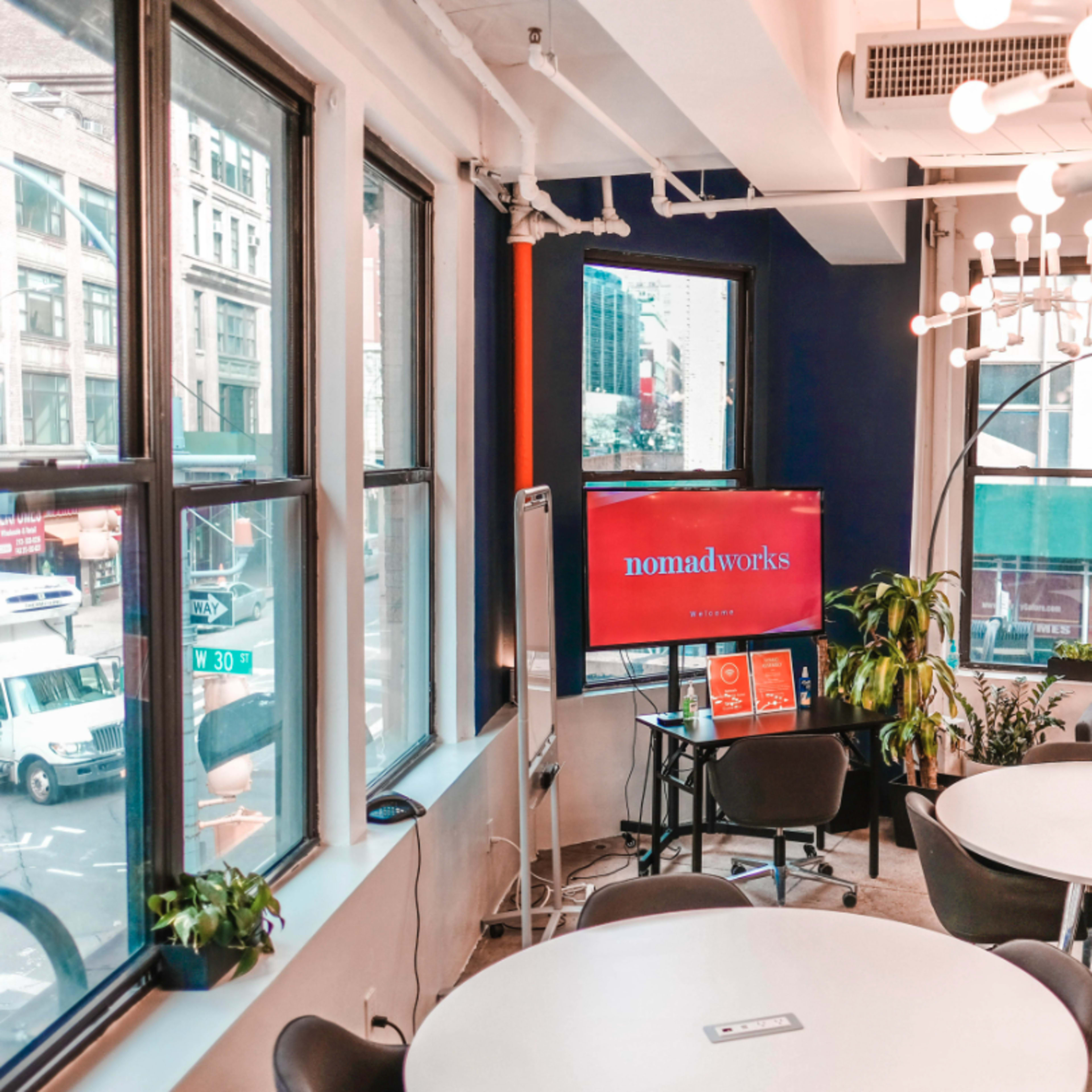 A modern coworking space with large windows, circular tables, a TV displaying "nomadworks," and a green plant in the corner.