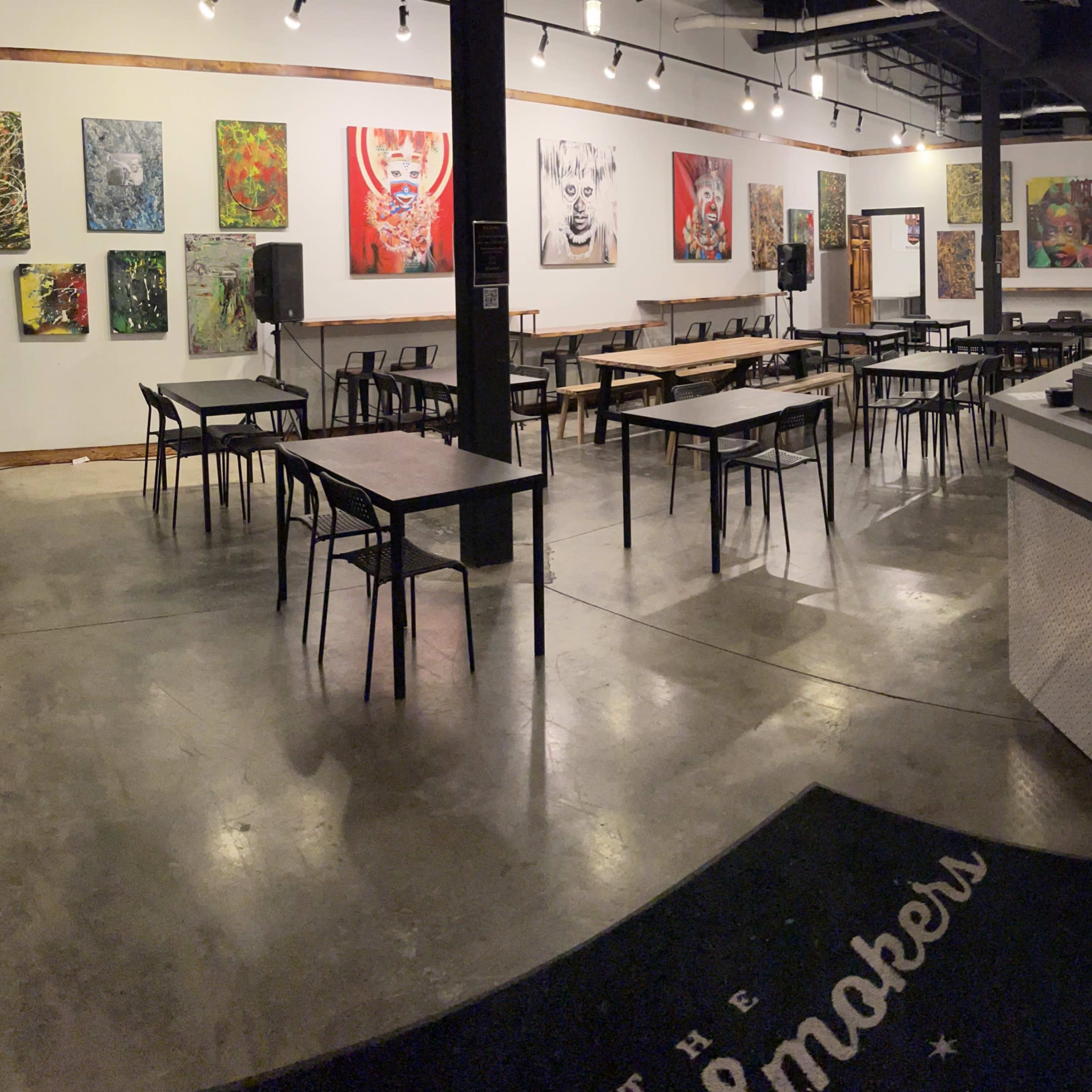 A spacious art gallery with vibrant paintings on the walls, several tables arranged across the floor, and a reception area in the corner.