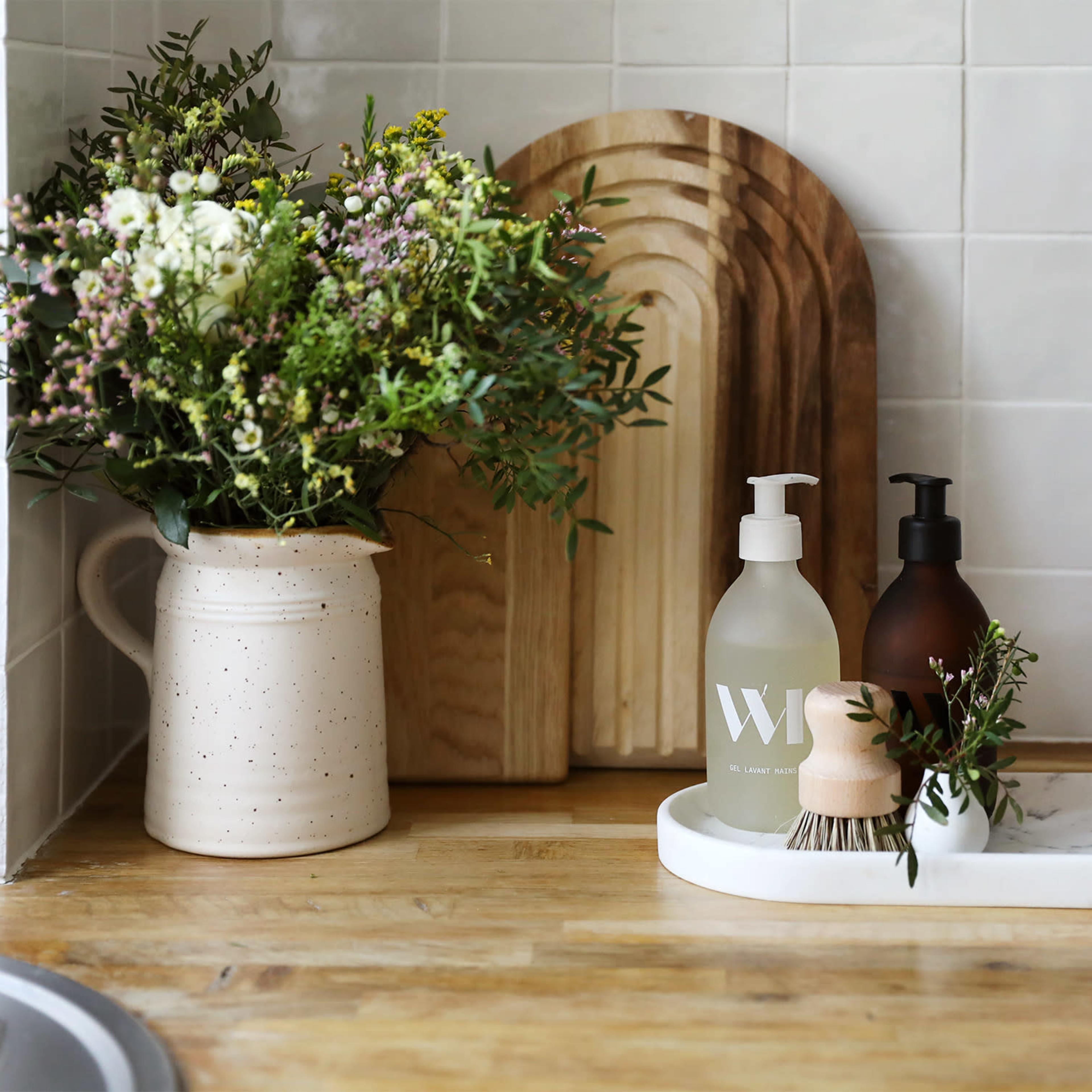 A wooden countertop displays a vase of flowers, two bottles of soap, and a small brush on a marble tray.