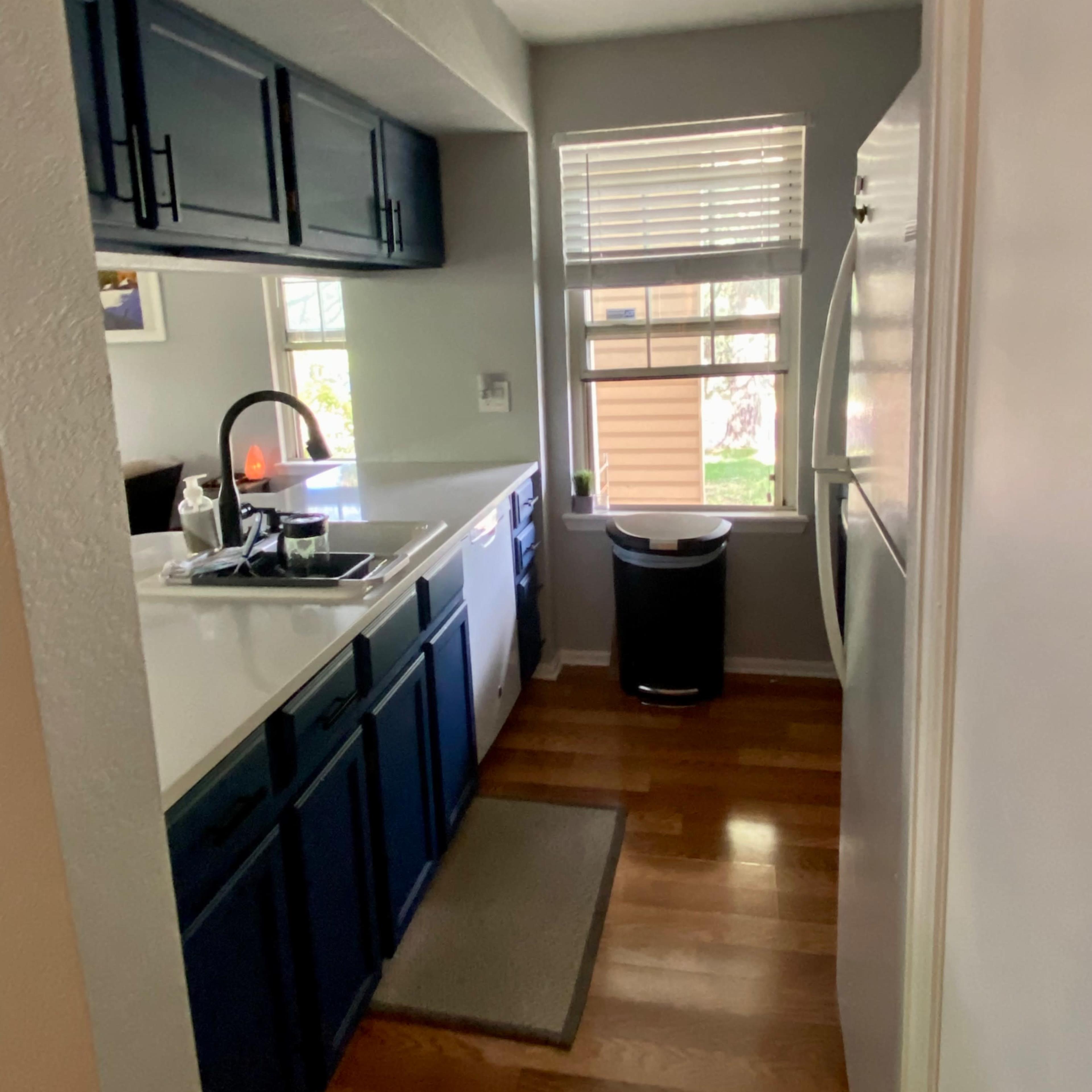 A narrow kitchen features blue cabinets, a white countertop, a window beside the sink, and a trash can near the refrigerator.