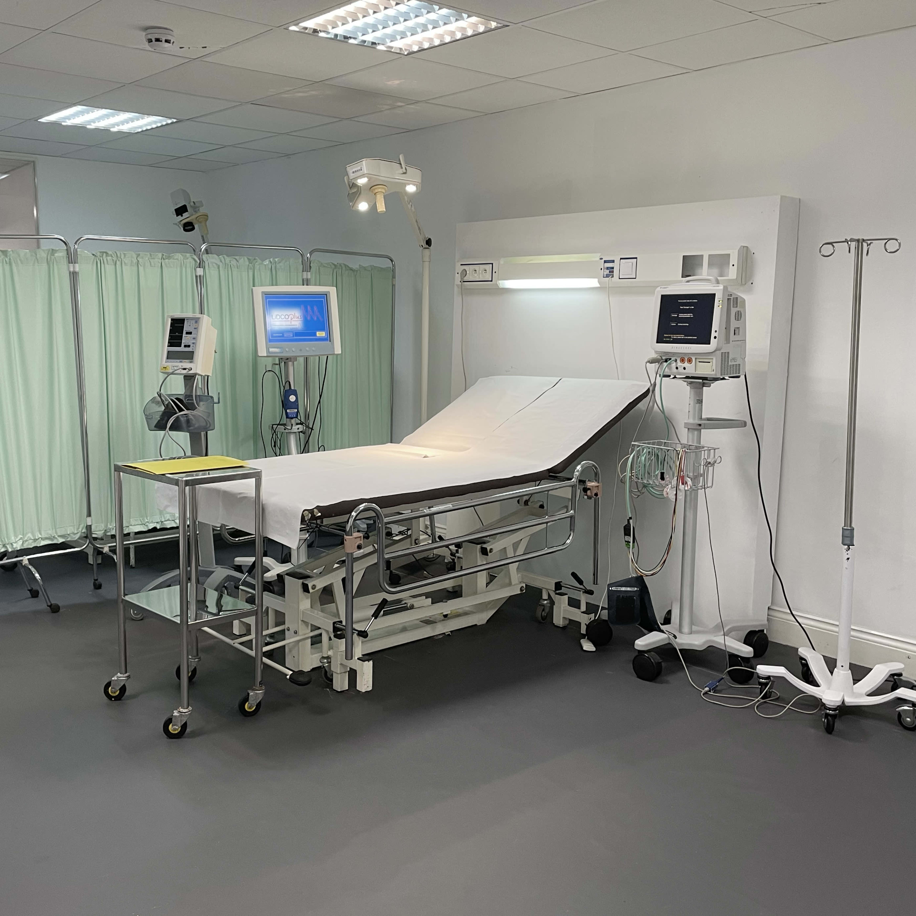 A medical examination room with an adjustable examination table, medical monitors, and green privacy curtains.