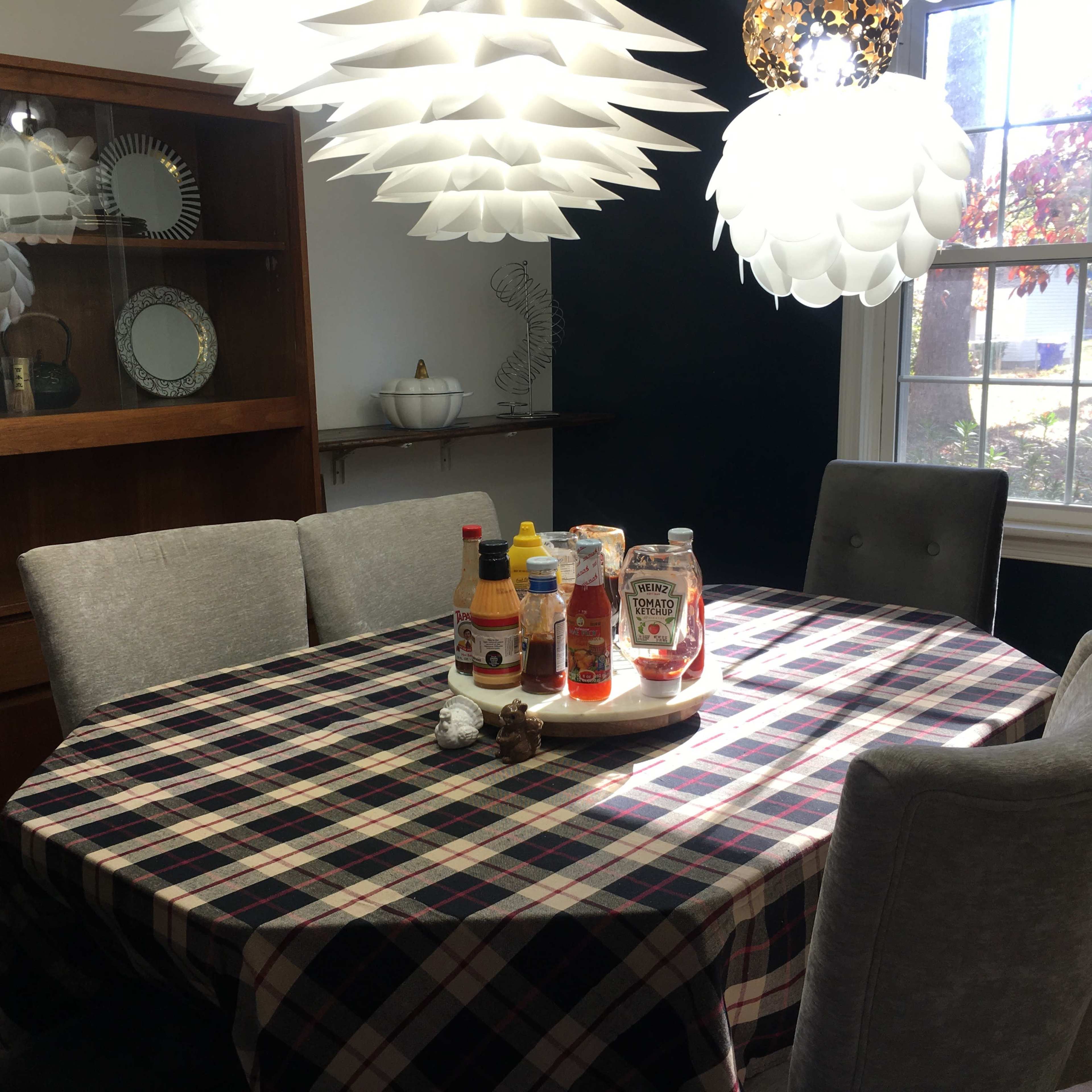 A round dining table with a plaid tablecloth is set with various condiments, surrounded by gray upholstered chairs, in a well-lit room featuring modern pendant lights and a wooden cabinet.