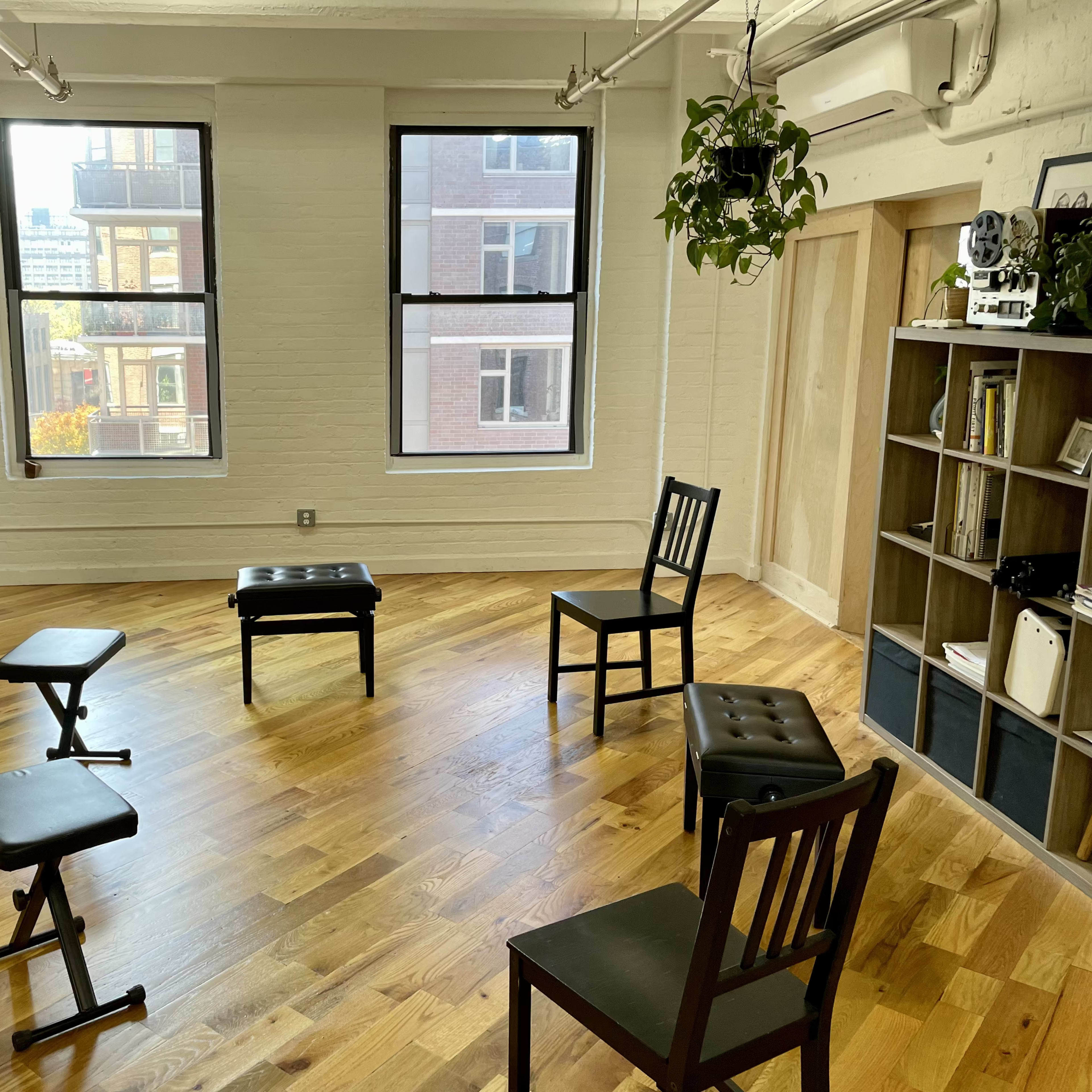 A sparsely furnished room with wooden floors, featuring a piano, several chairs, and decorative hanging plants.