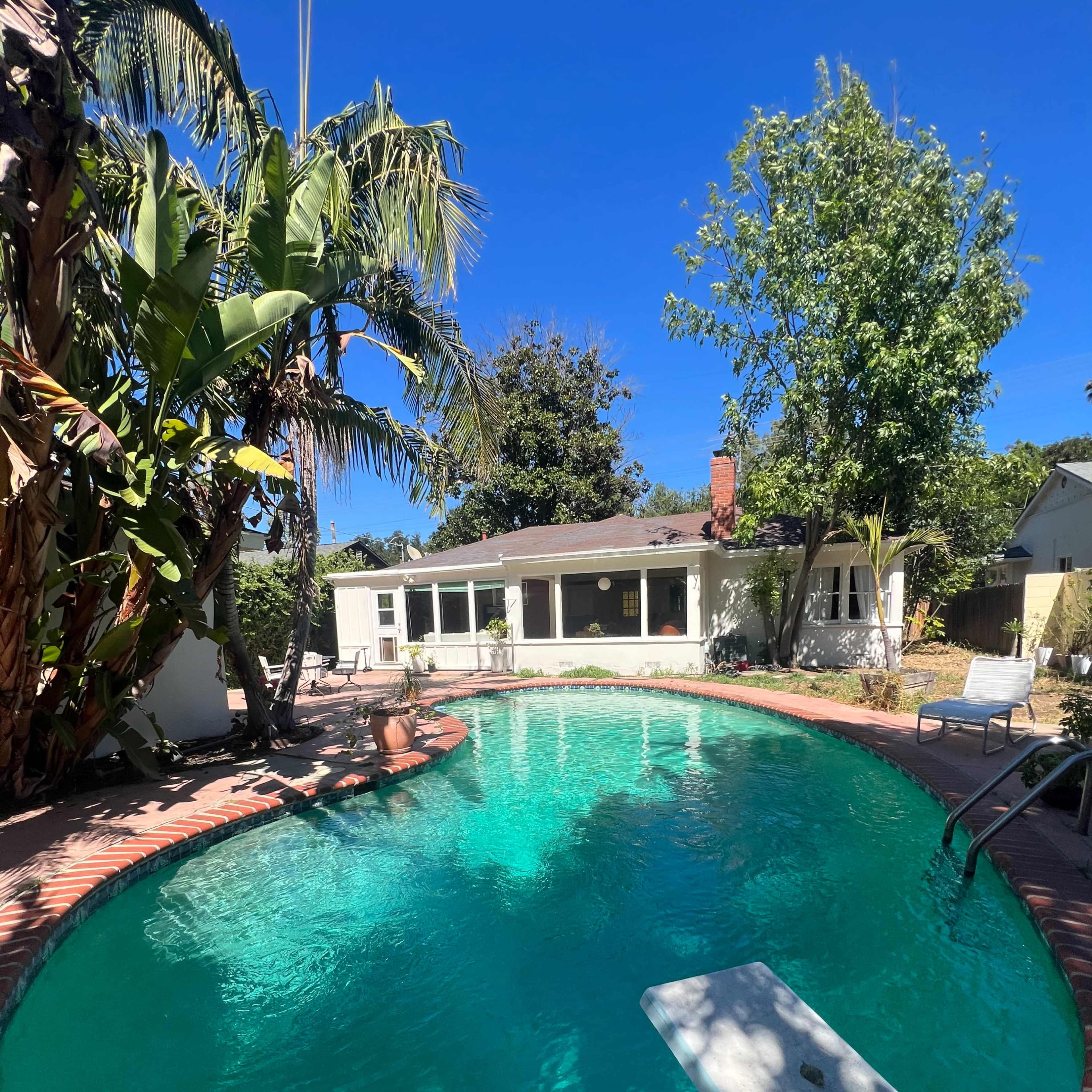 A circular swimming pool is surrounded by a patio, palm trees, and a house in a sunny outdoor setting.