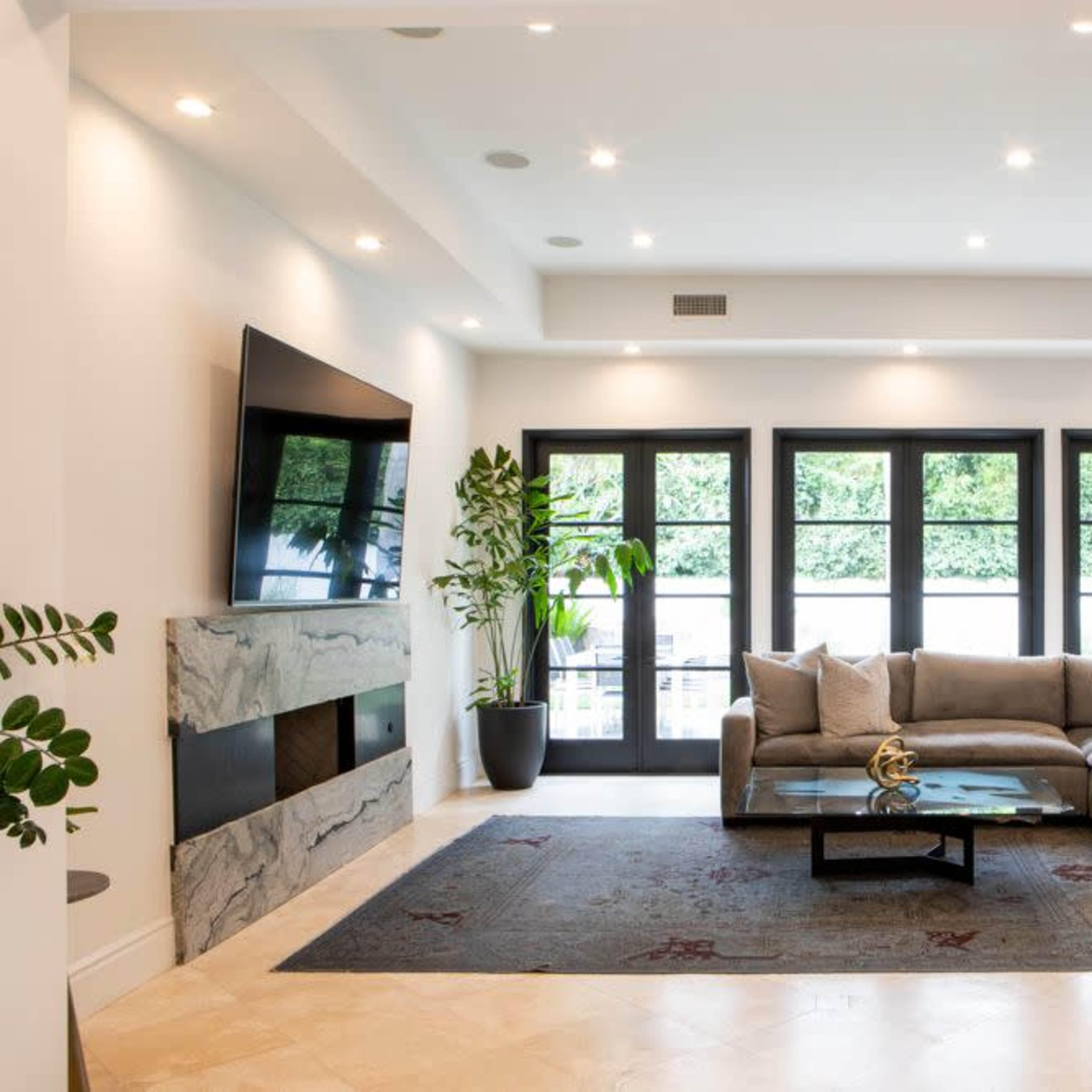 A modern living room with a large gray sectional sofa, a marble feature wall, a flat-screen TV, and large black-framed windows.