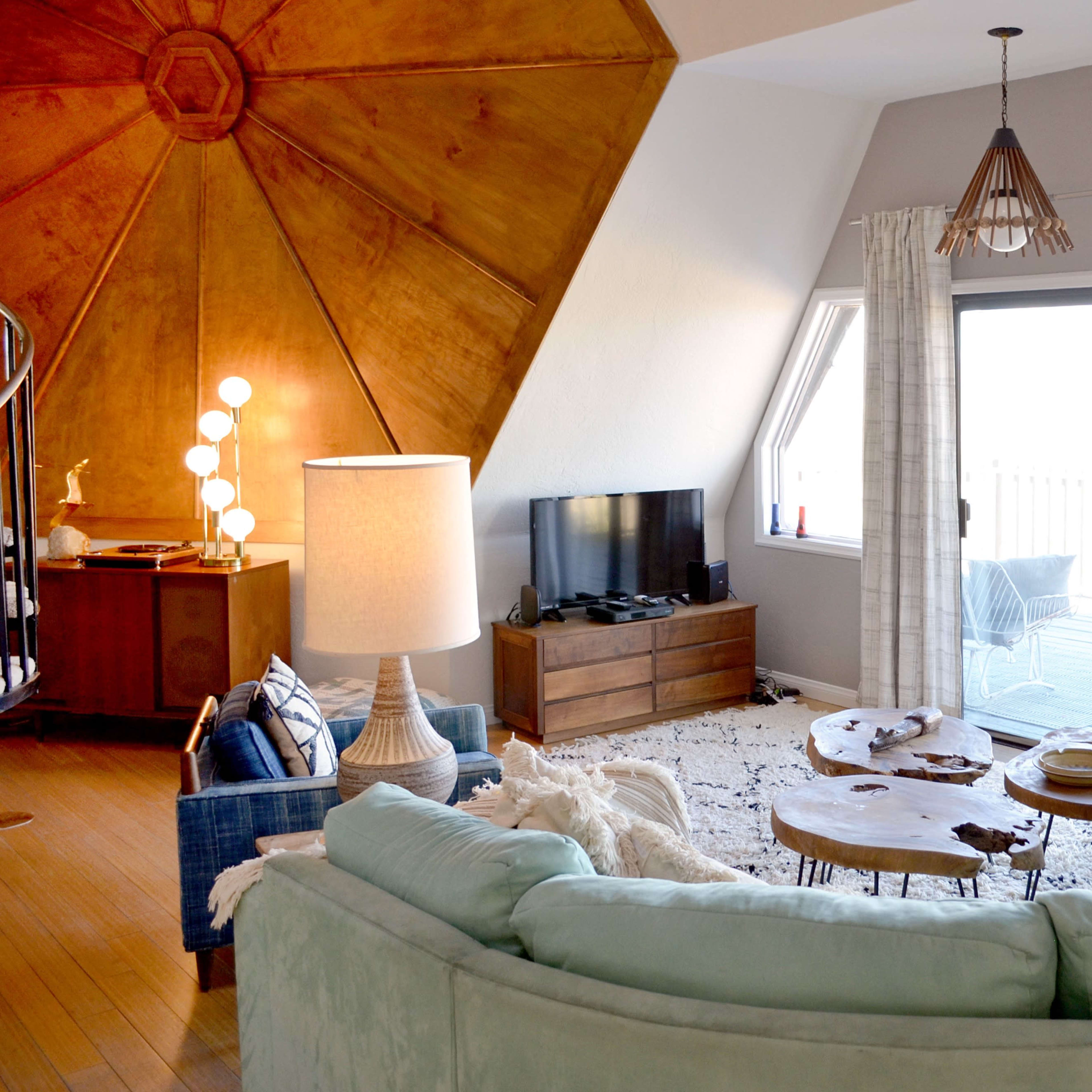A circular living room features a light blue sectional sofa, two wood coffee tables, a large television, and wide windows overlooking a deck.