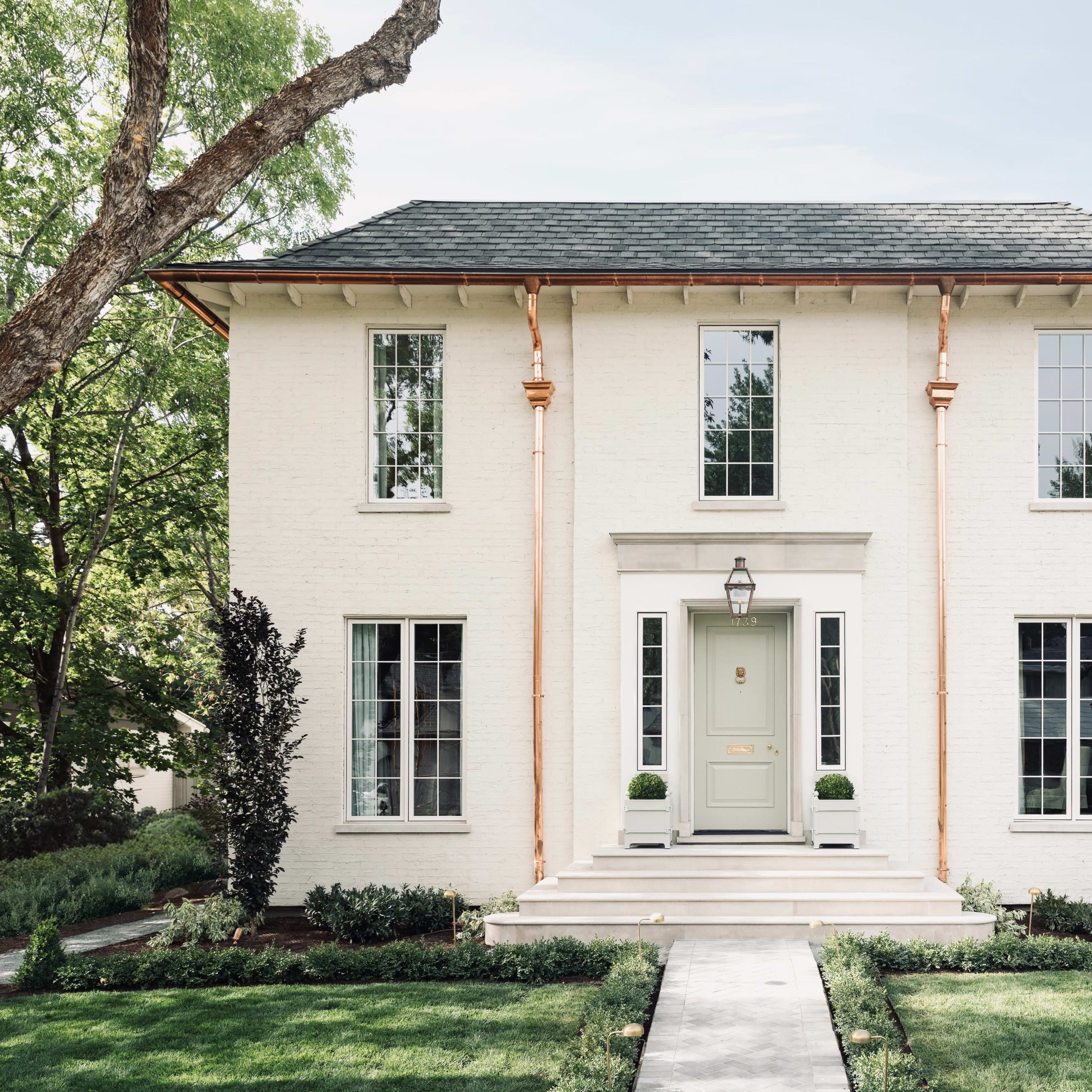 A two-story white brick house with large windows, a central front door, and a landscaped yard featuring symmetrical greenery and a concrete walkway.