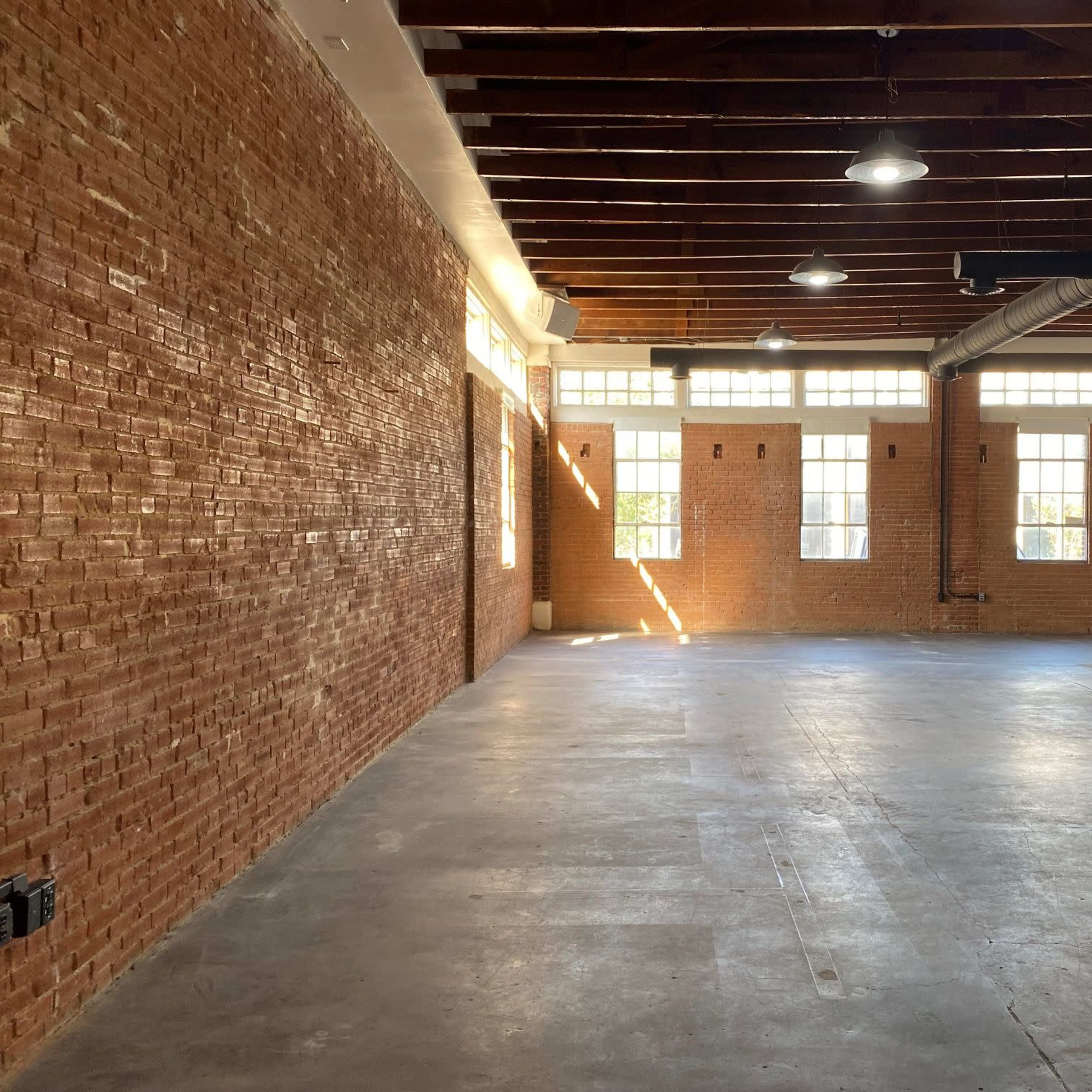 A spacious room with exposed brick walls, large windows, and a concrete floor.