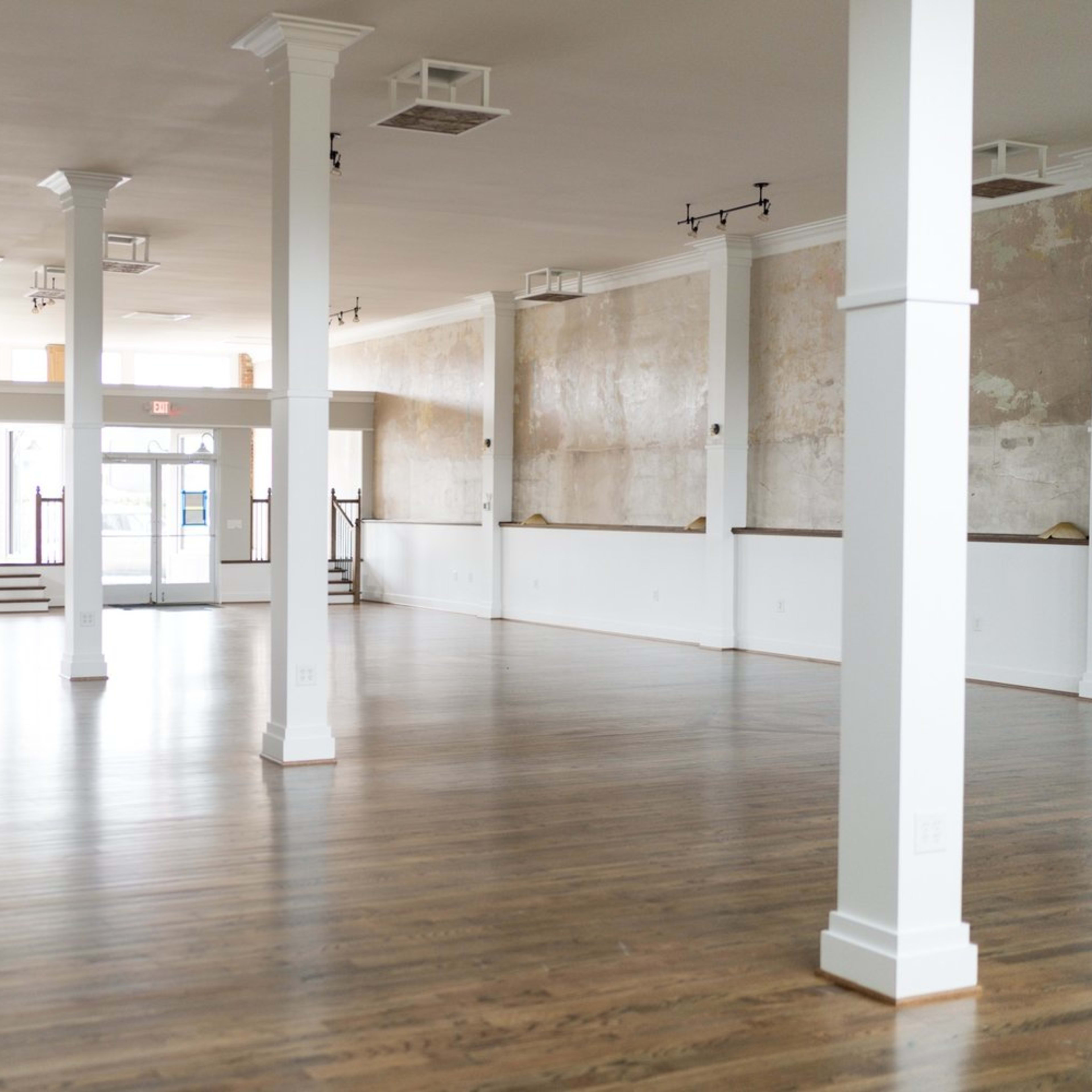 A spacious, empty hall with polished wooden floors, tall white pillars, and large windows letting in natural light.