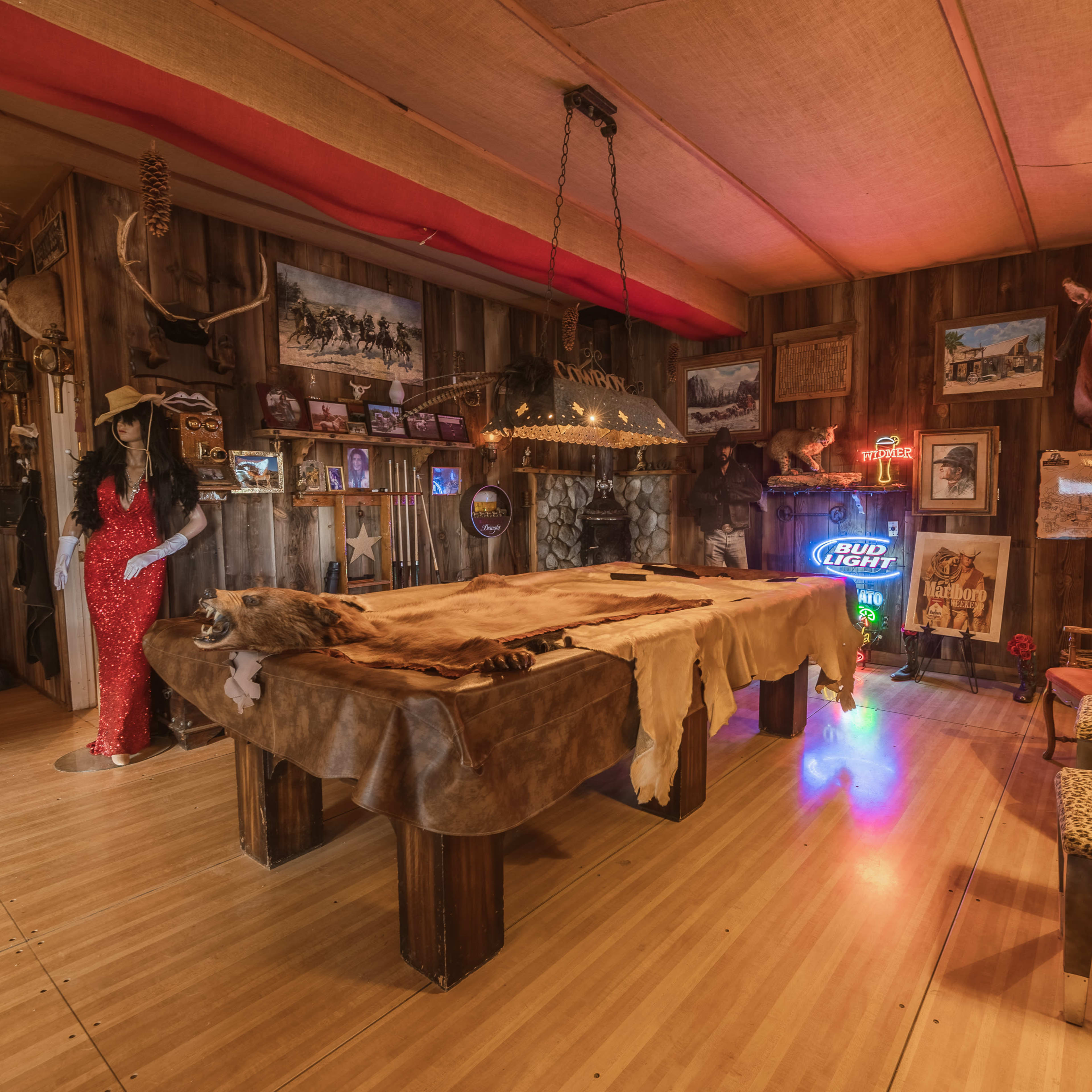 The interior of a rustic room features a large wooden table covered with animal hides, surrounded by decorative chairs and adorned with various mounted animal heads and framed photographs on the walls.