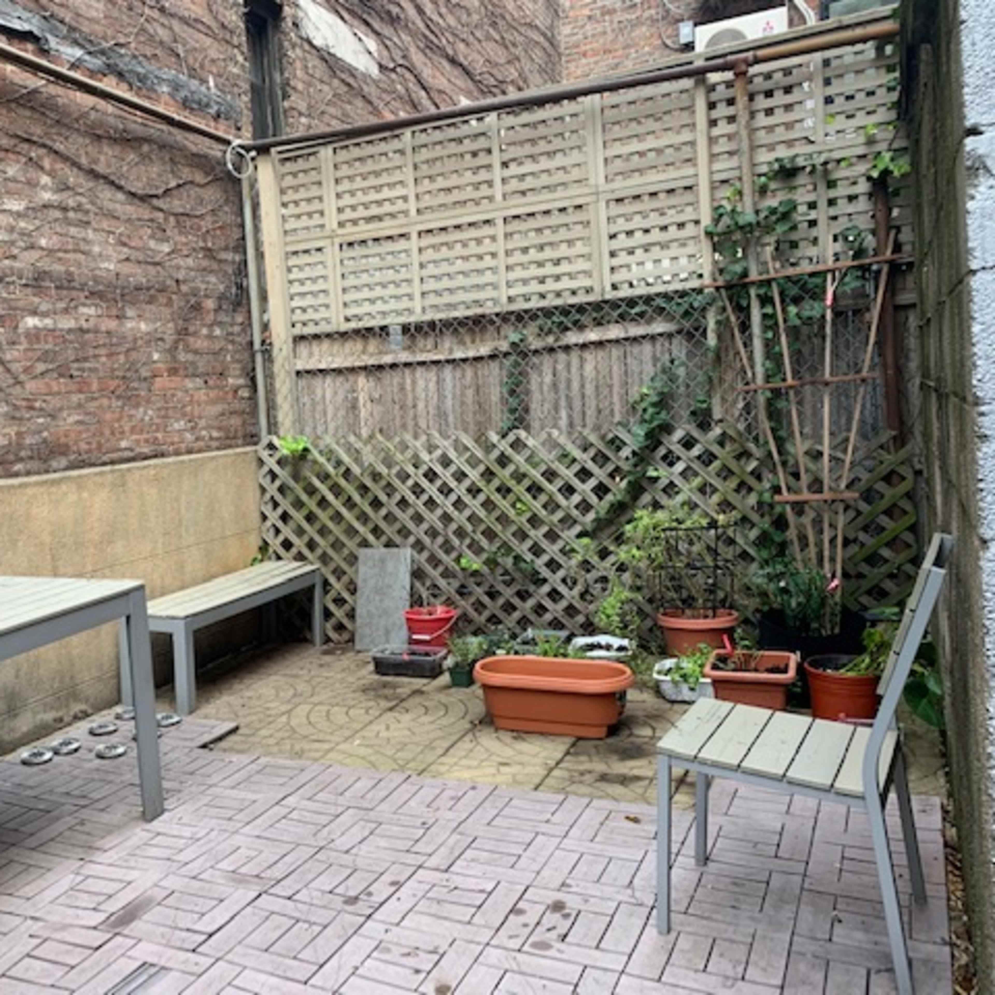 A small outdoor patio area with two wooden tables, several potted plants, and a trellis against a brick wall.