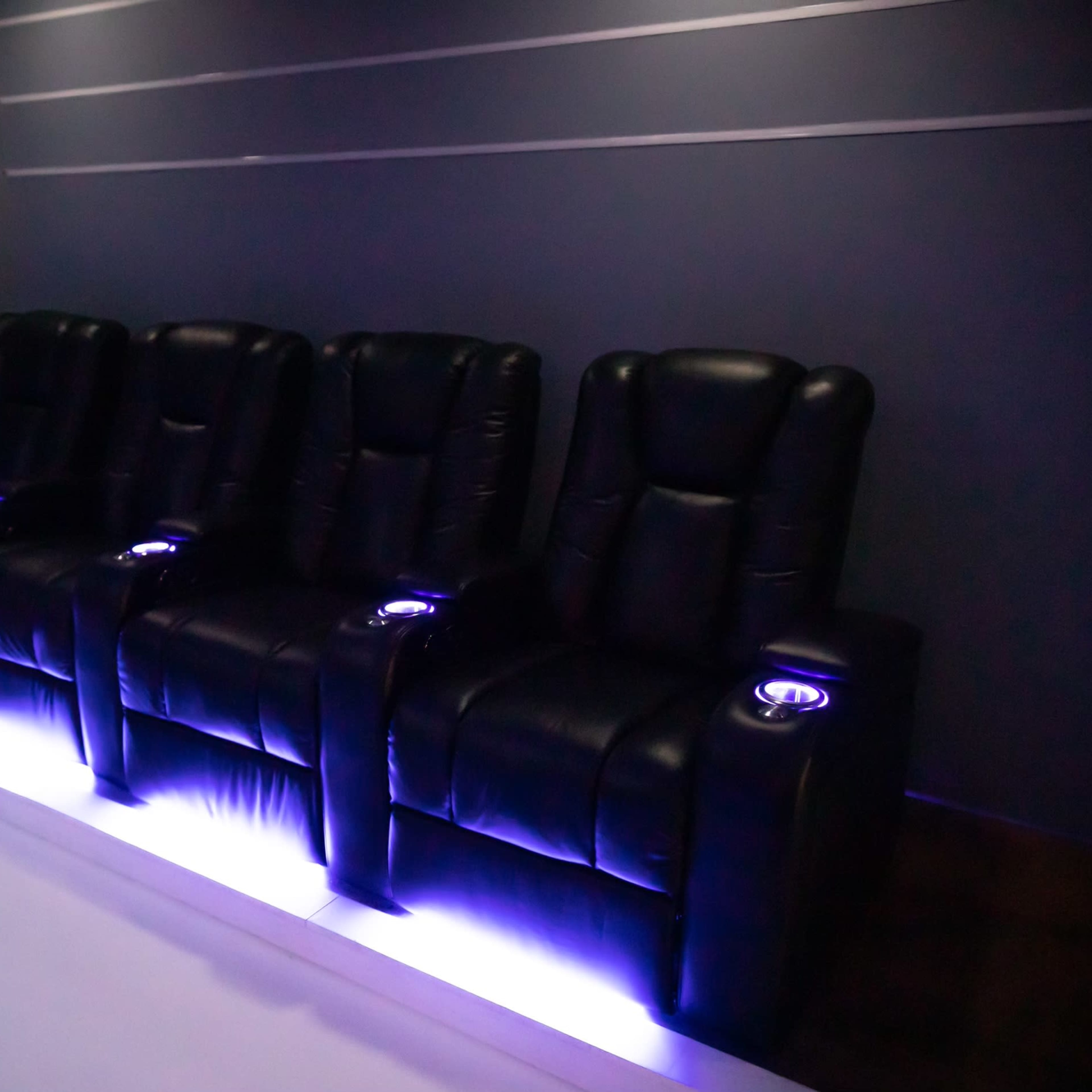 A row of four black leather reclining chairs with under-seat LED lighting in a dimly lit room.