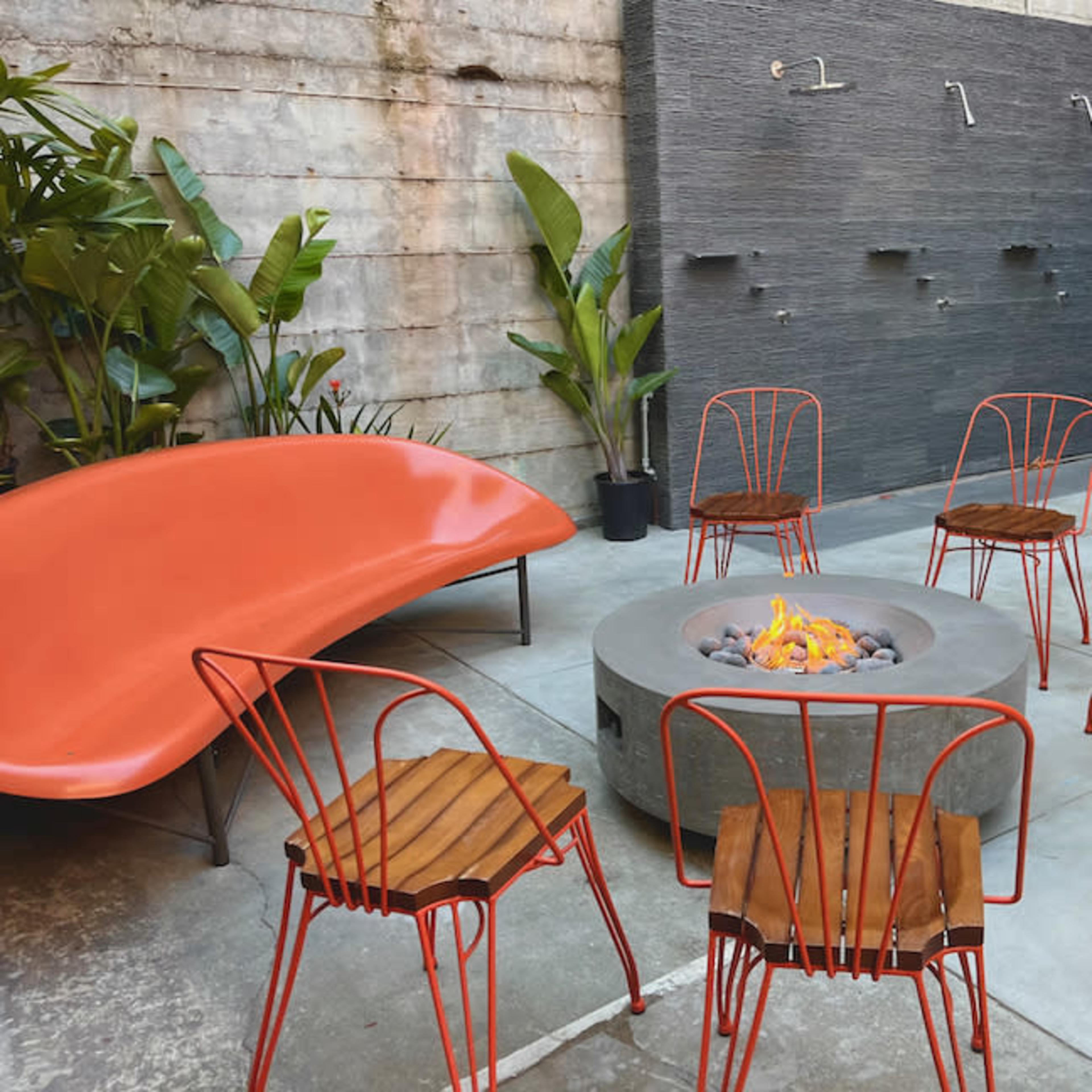 A modern courtyard with a circular fire pit surrounded by six wooden and metal chairs, a distinctive orange lounge chair, and a textured wall with water fixtures.