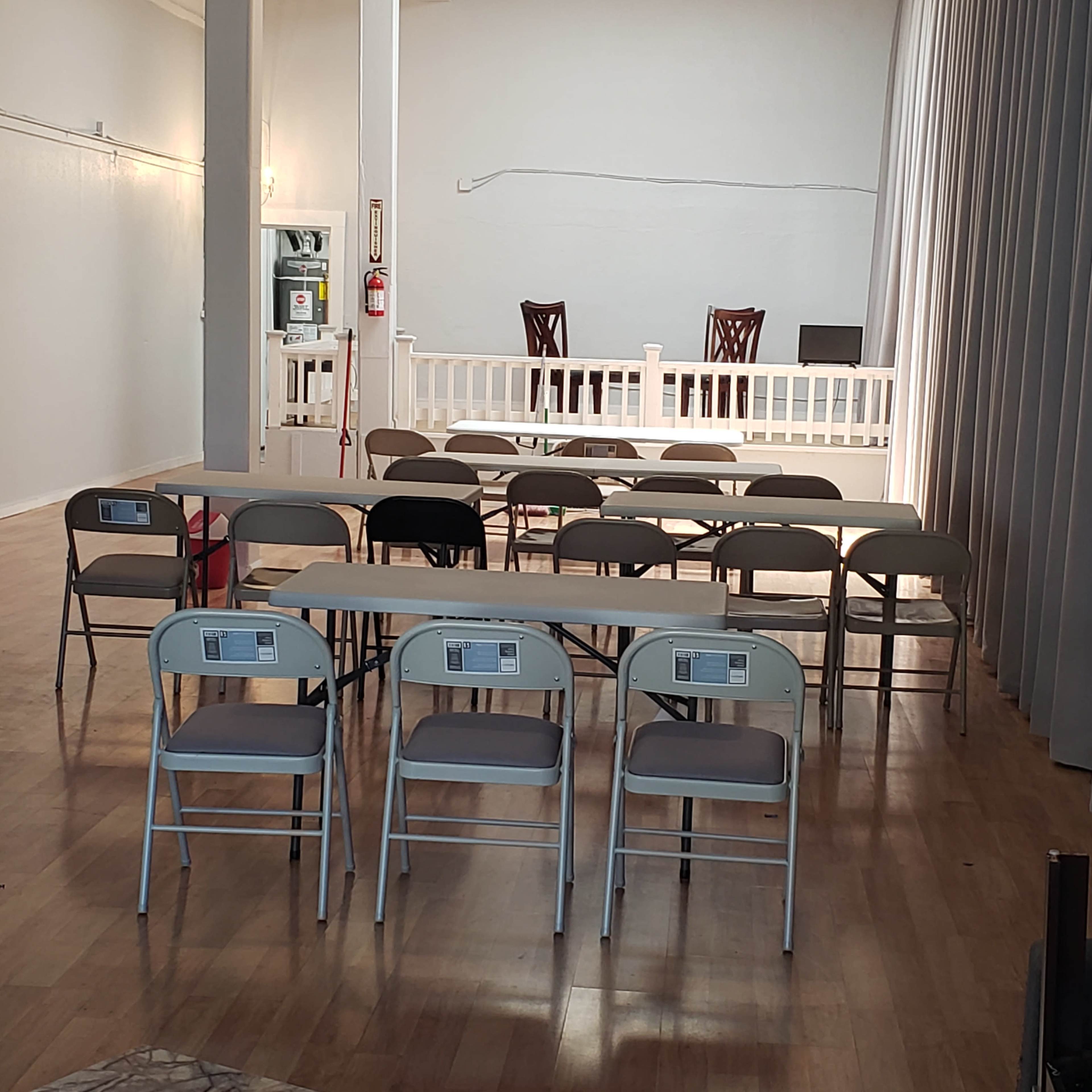 A spacious room with rows of tables and chairs facing a small stage area, flanked by curtains.