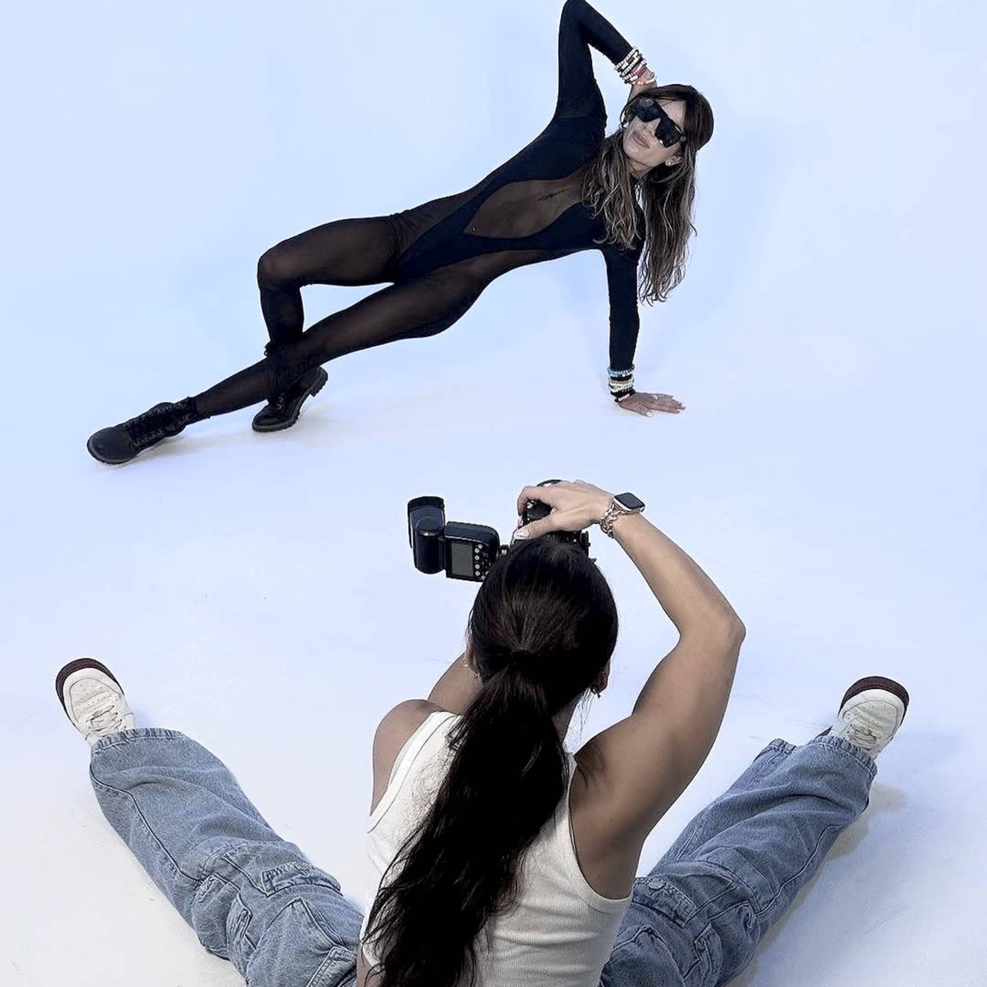 A model poses in a black bodysuit on a white backdrop while a photographer captures the shot from a seated position.