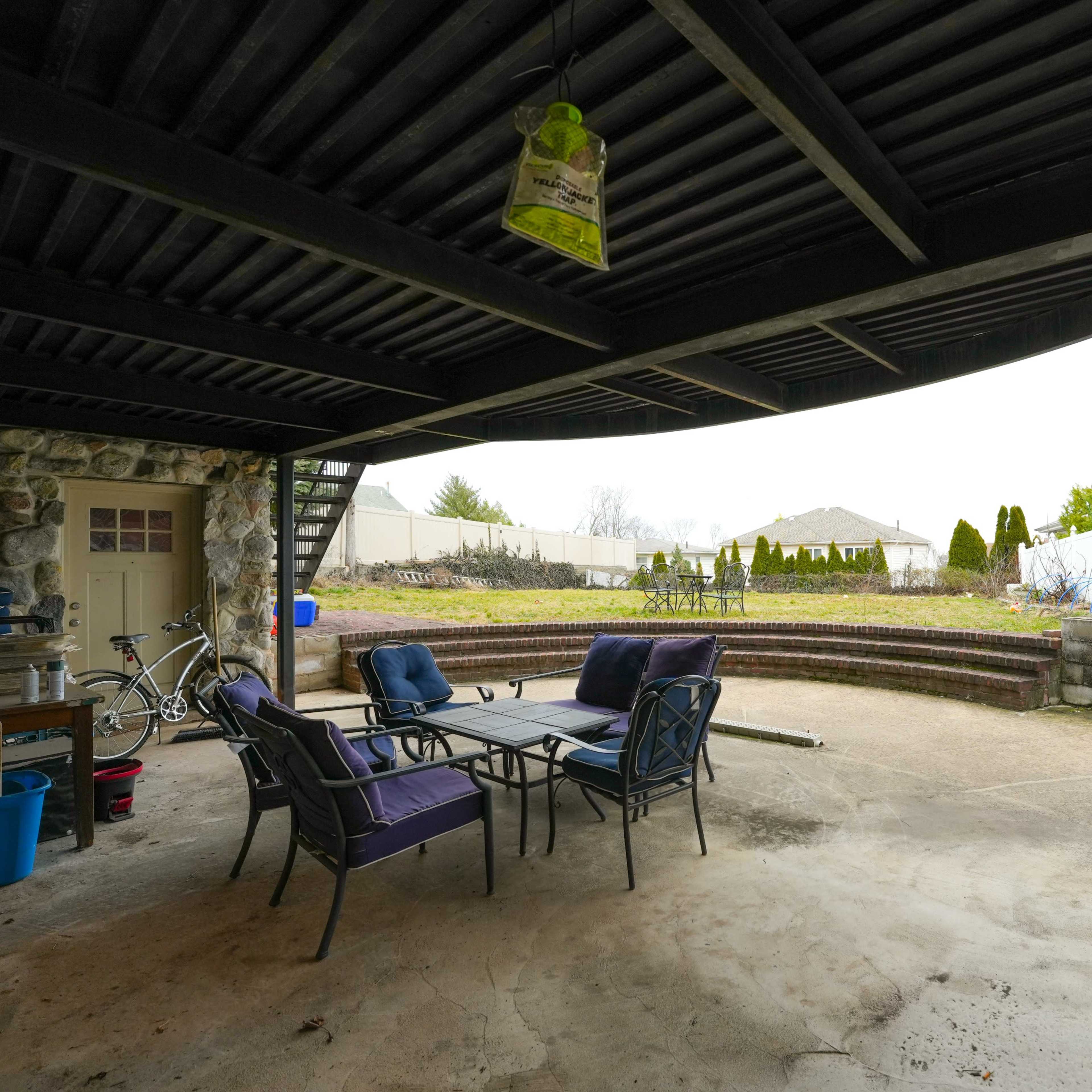 A covered outdoor patio area with a table and chairs, a barbecue grill, and a view of a garden and landscaping in the background.