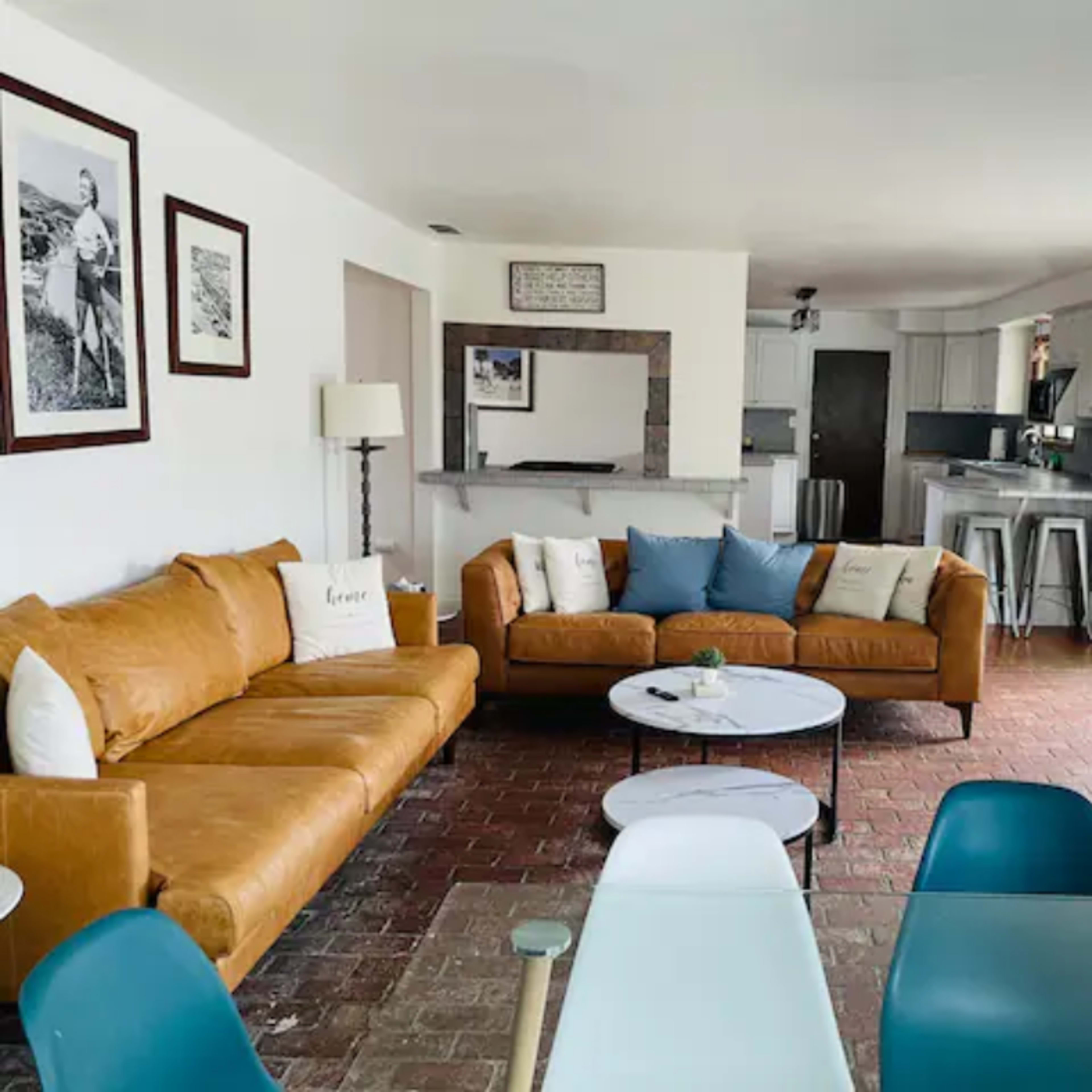 A living room with a brown sectional sofa, a round coffee table, and a dining area featuring modern chairs and a kitchen in the background.