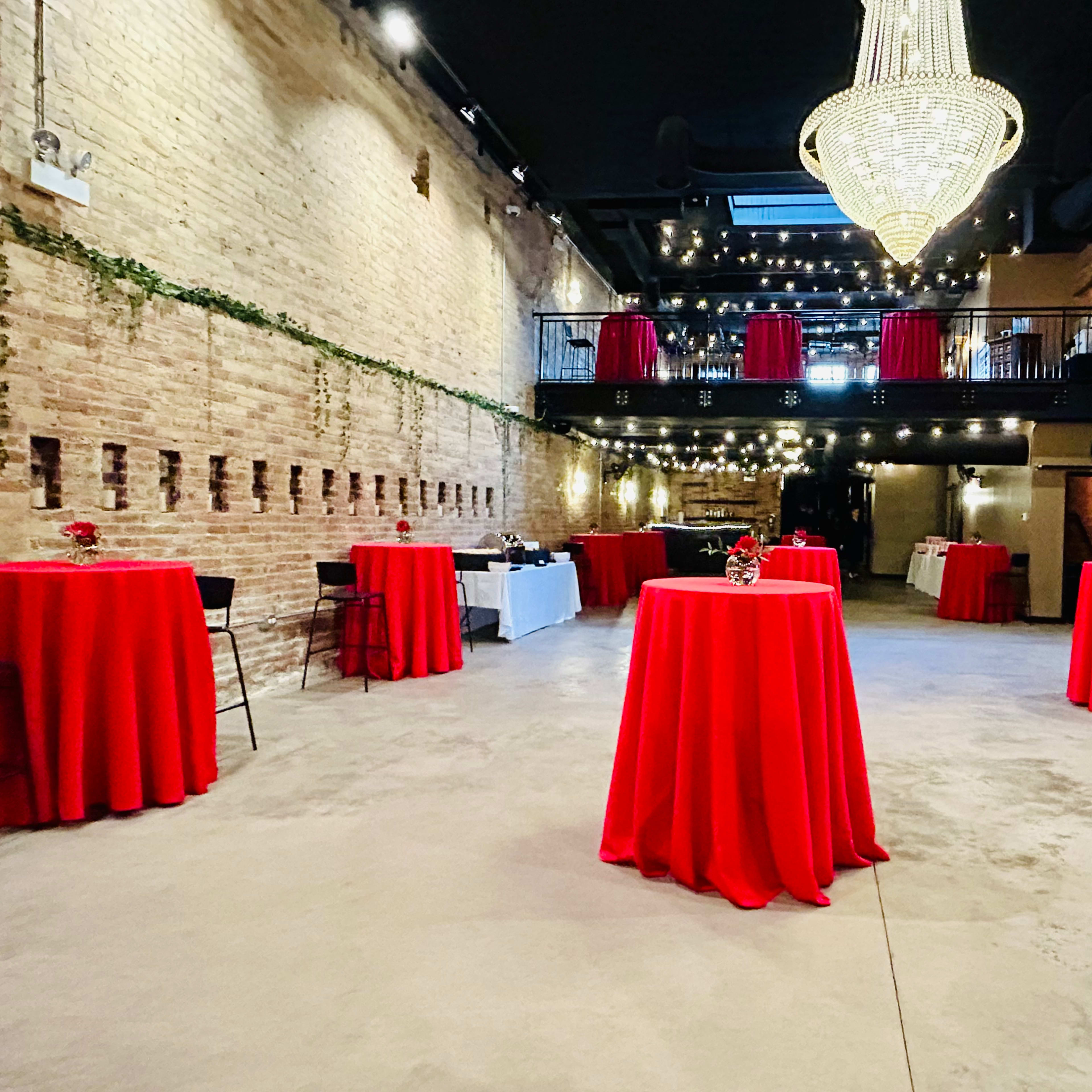 The spacious venue features red tablecloths draped over tall tables, with a chandelier hanging from the ceiling and exposed brick walls.