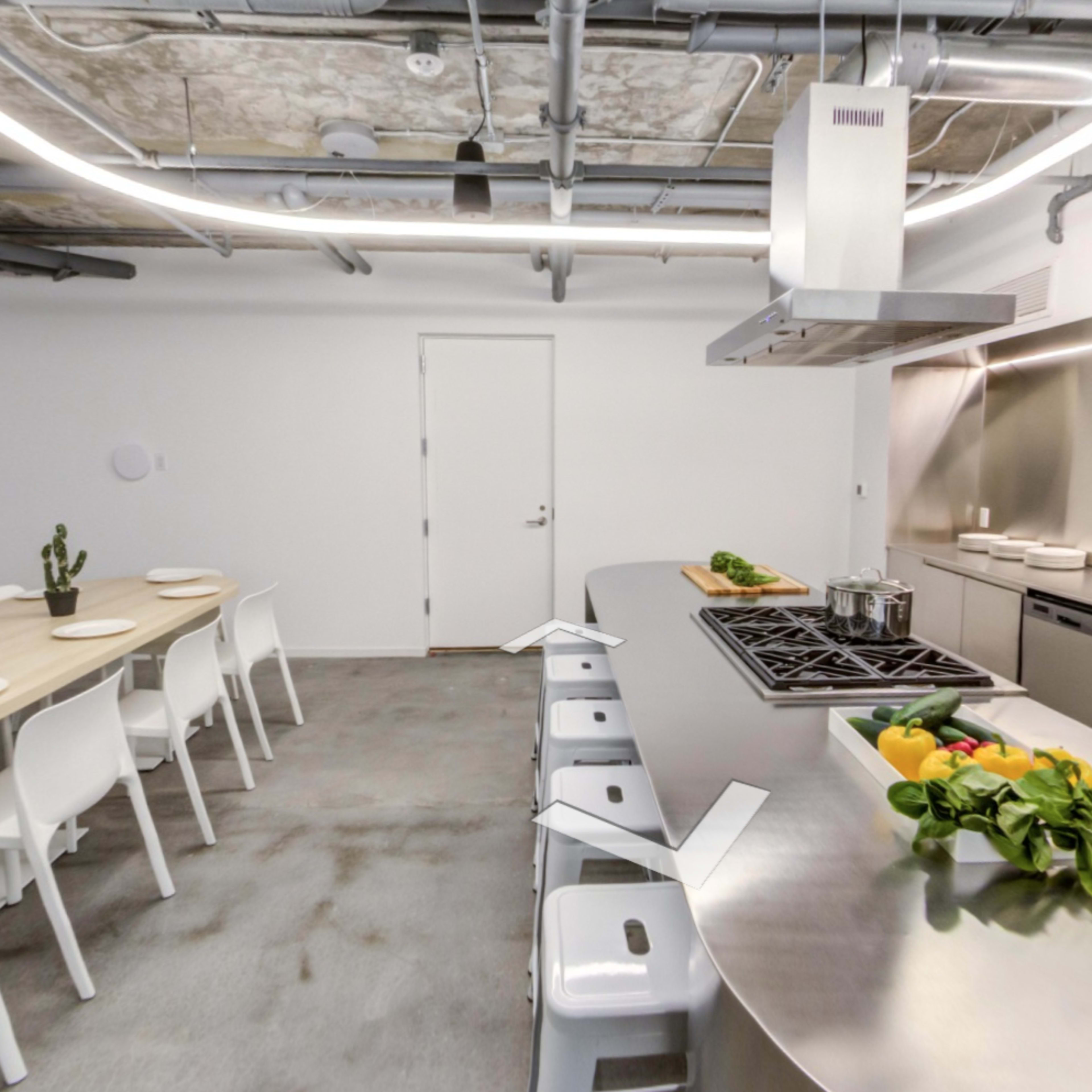 A modern kitchen features a long wooden table set for a meal, metal stools, and a stainless steel cooking area with a hood and stovetop.