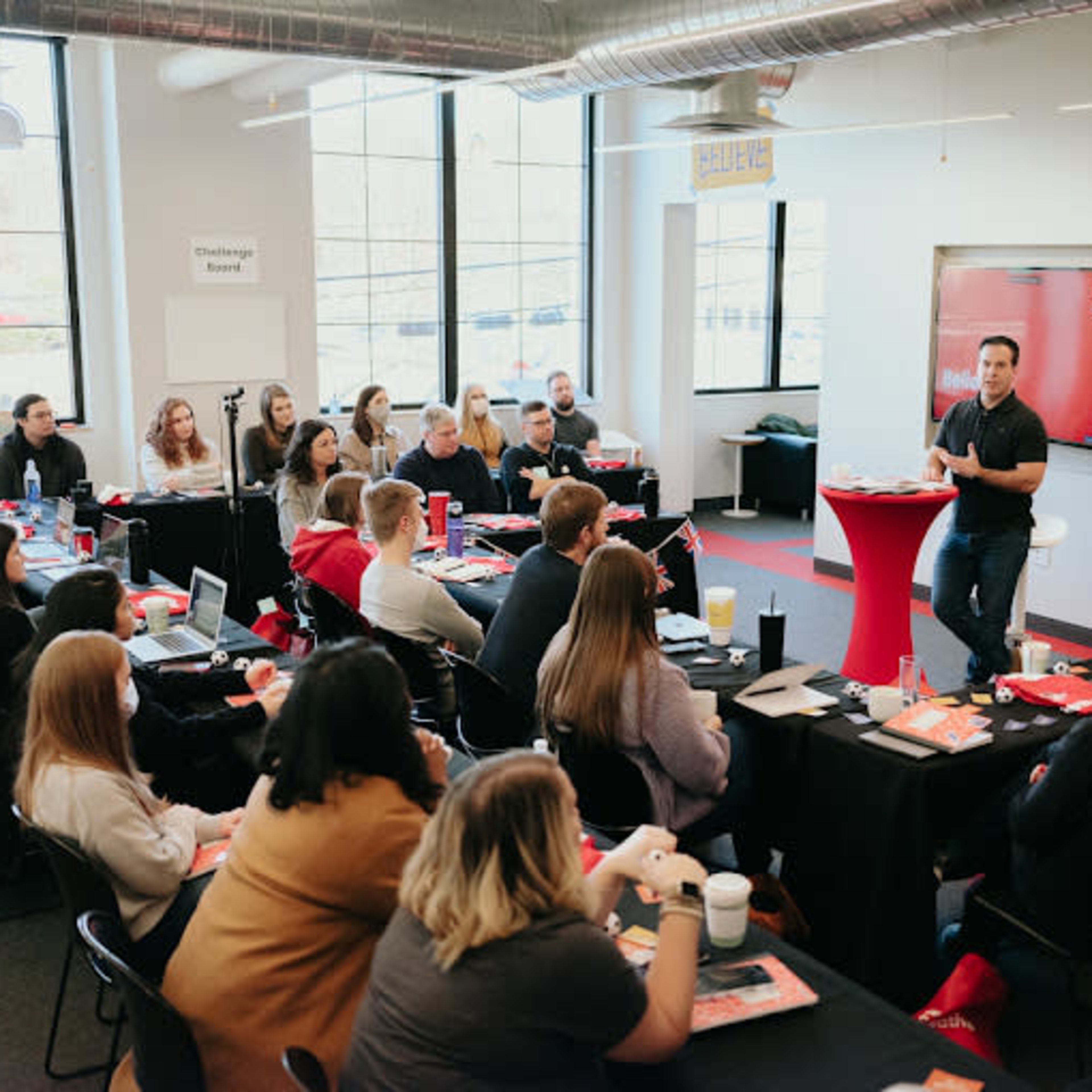 A speaker stands in front of an audience seated at tables in a conference room, presenting on a screen that displays the word "Believe."